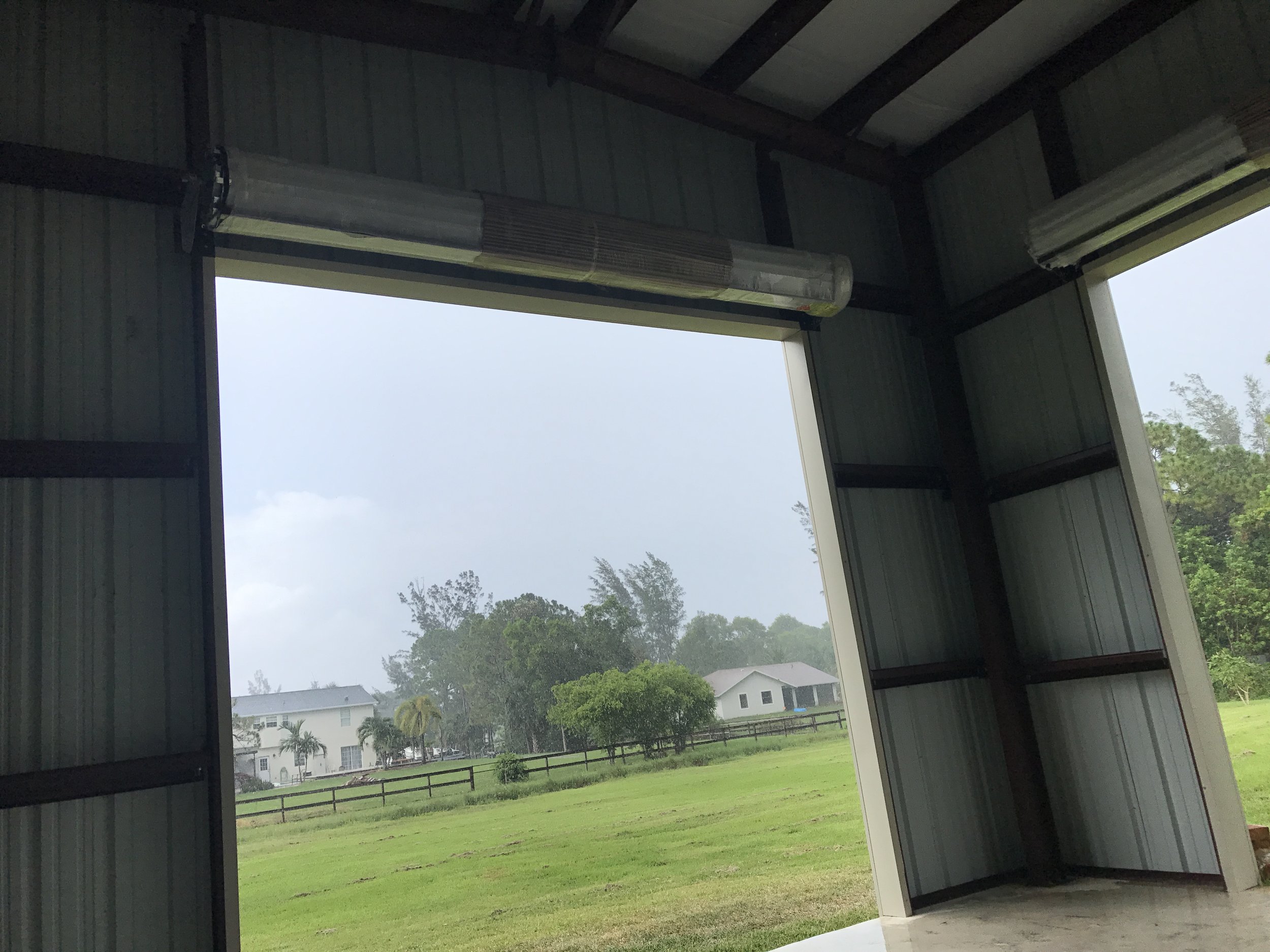 View from inside an open metal building looking out onto a lush green yard with trees and houses in the distance on a cloudy day.