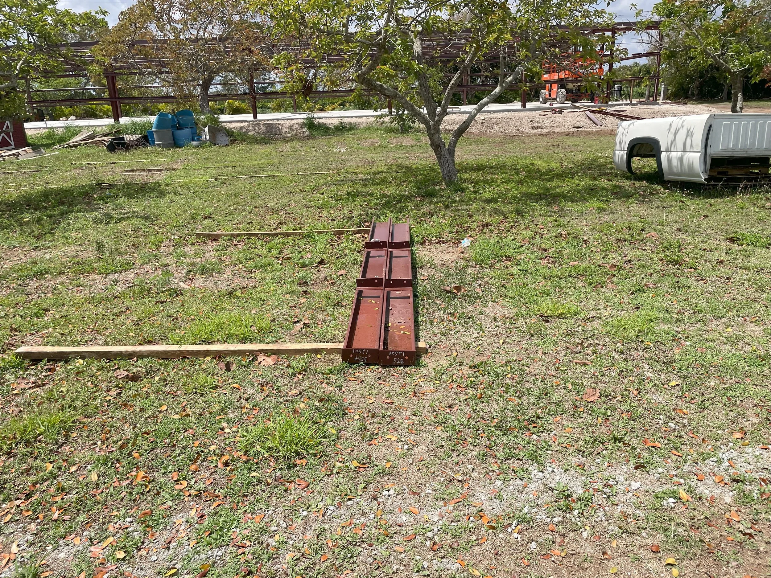A grassy lot with a tree in the center, construction materials, and a partially assembled metal structure. Blue containers and a white truck bed are nearby.