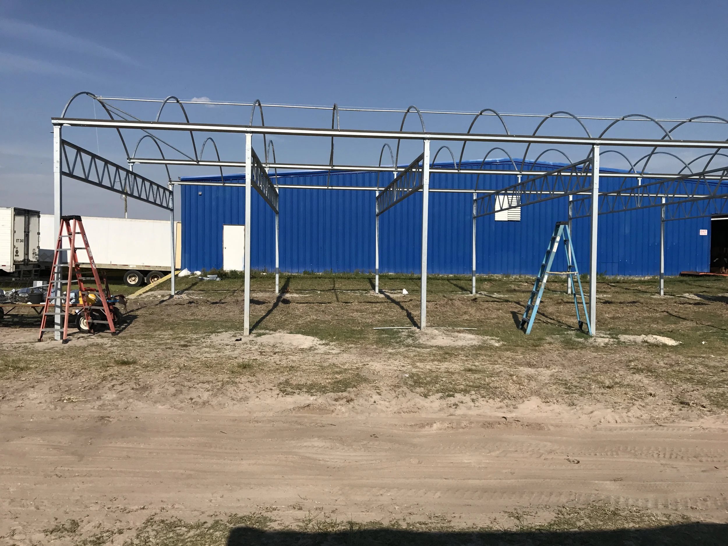 Metal framework structure erected in front of a blue building with ladders nearby, on a dirt and grass lot under a clear sky.
