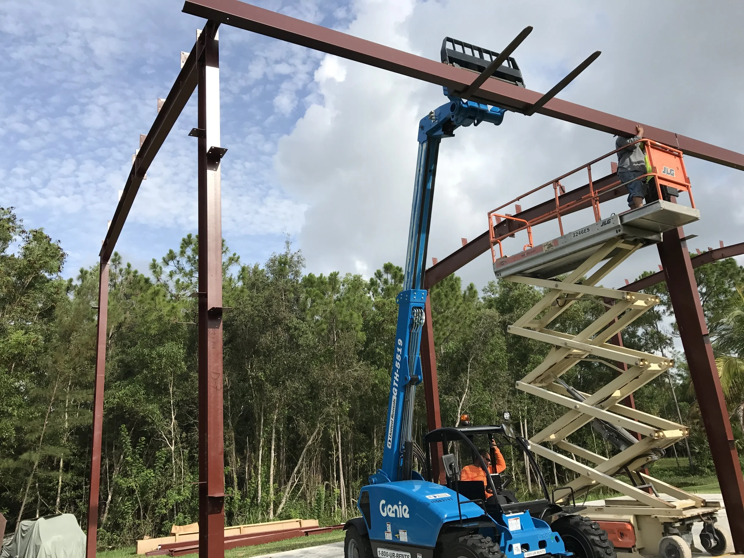 Construction workers building a steel frame structure using a scissor lift and crane in an outdoor setting with trees and blue sky.