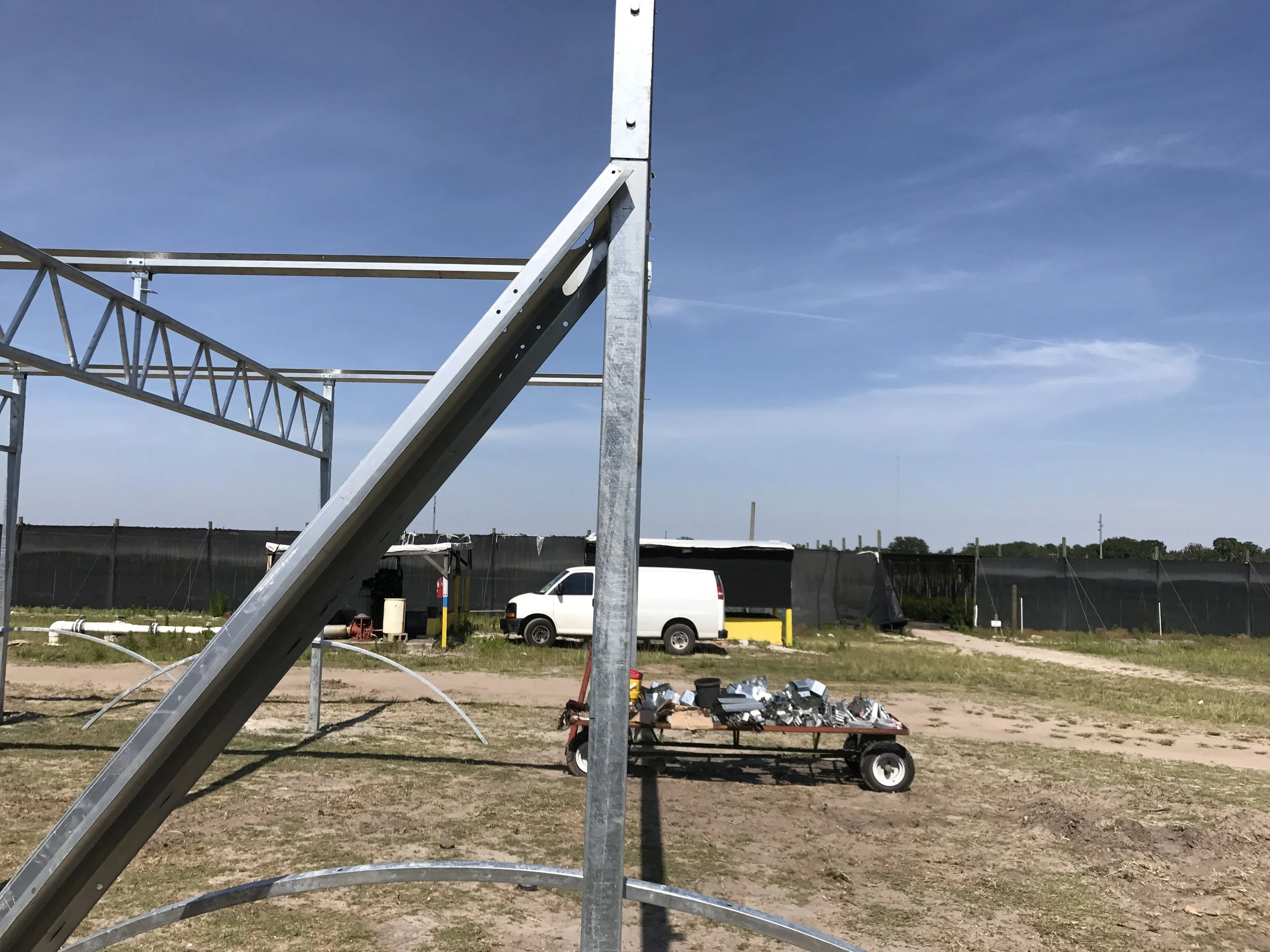 Empty outdoor construction site with metal framework, a cart filled with tools, a white van, and a black fence under a blue sky.