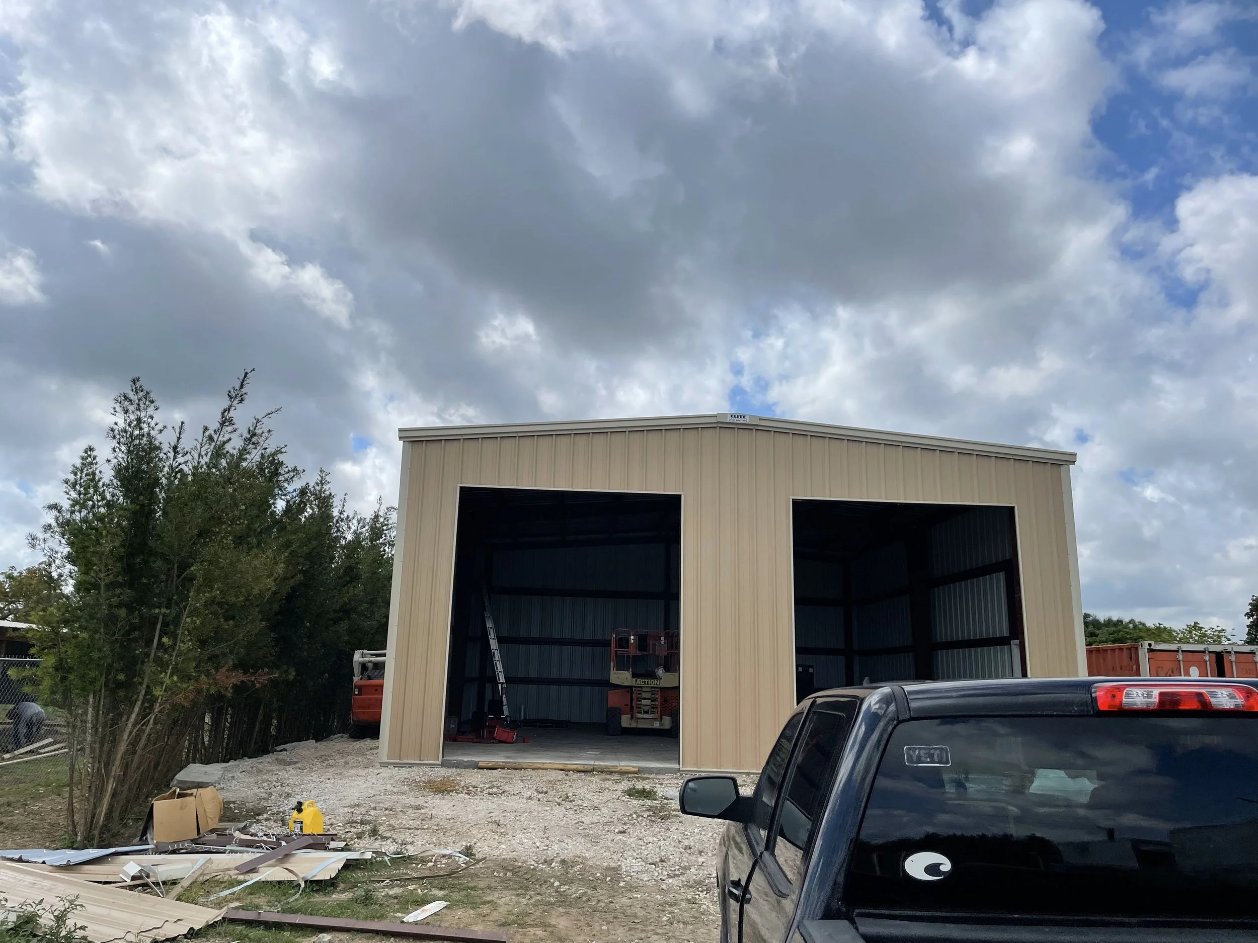 A metal building under construction with two large open bays, a black pickup truck parked in front, and construction tools and materials scattered on the ground. There are trees and a cloudy sky in the background.