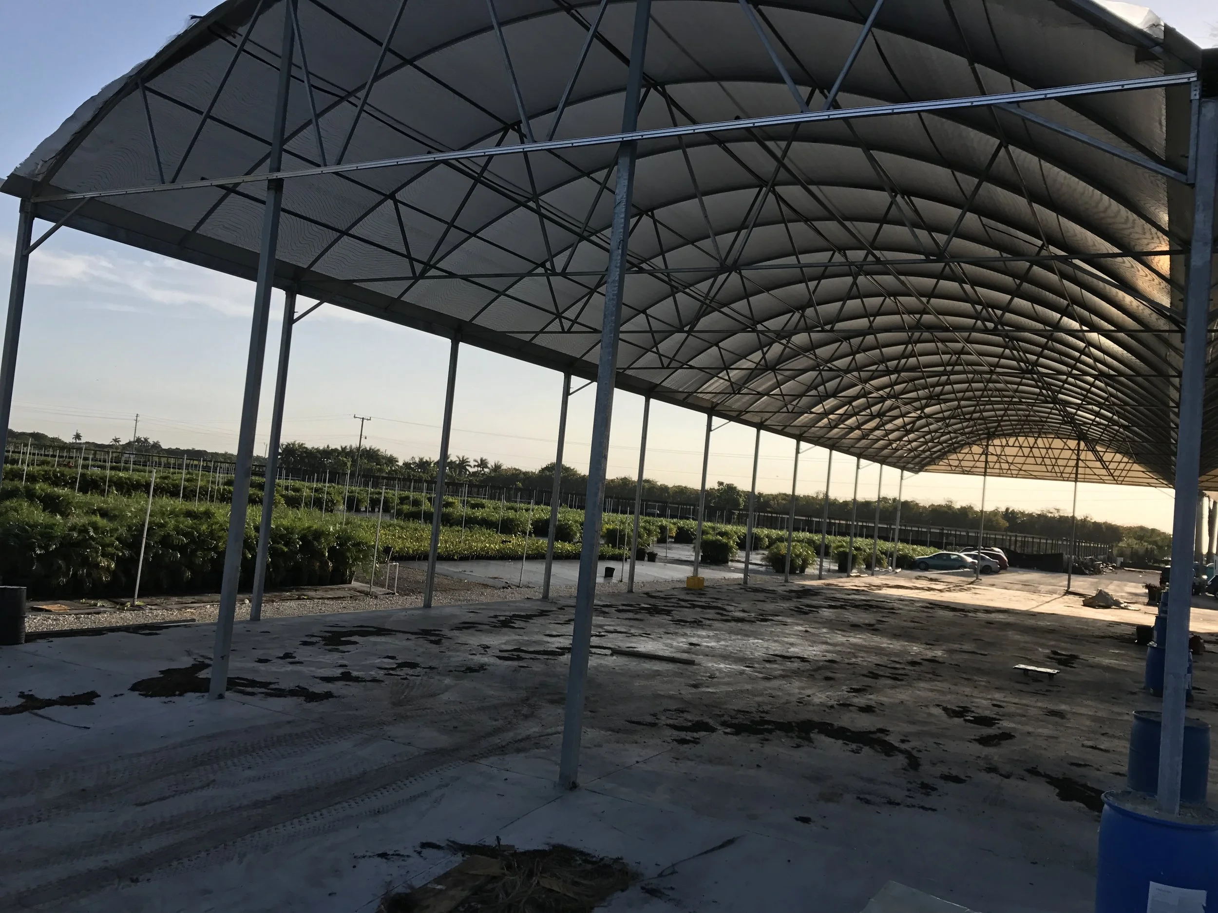 A large outdoor agricultural structure with a metal frame and a curved roof, situated over a dirt and concrete area, with green fields and parked cars in the background.