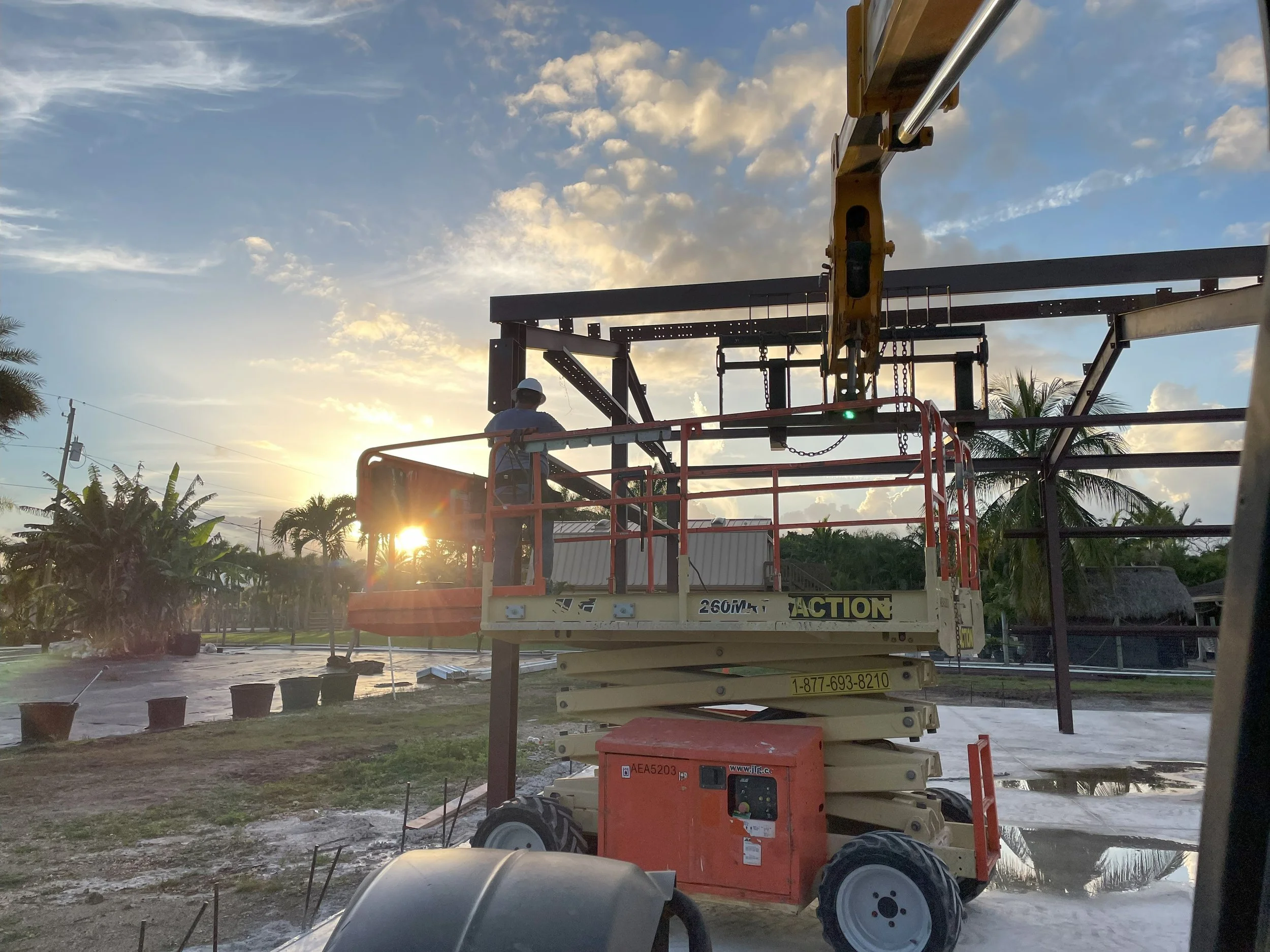 A construction worker on a scissor lift working on a steel framework at sunset, with trees and a partly cloudy sky in the background.