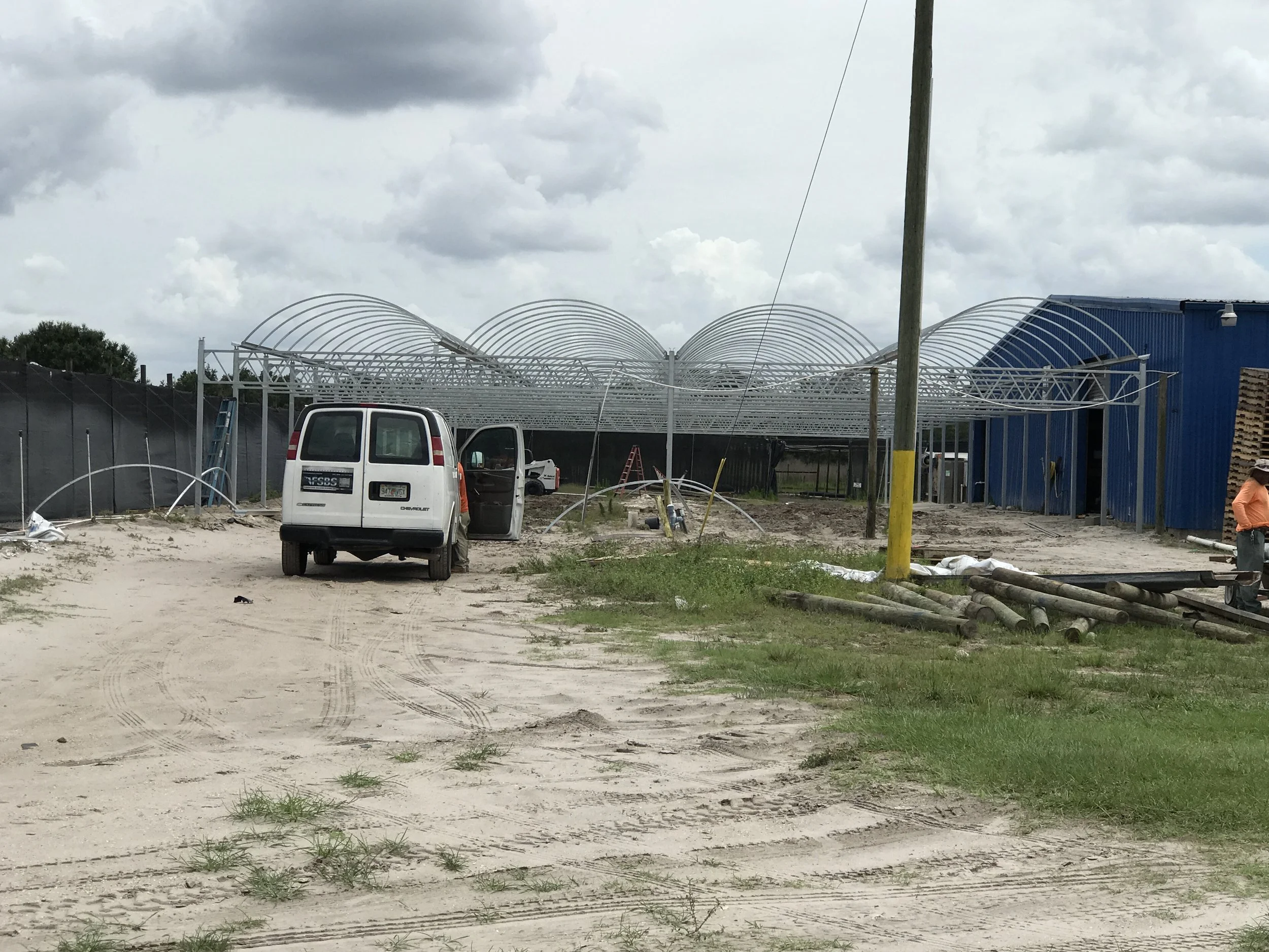 Construction site with a vehicle and workers building a large metal structure adjacent to a blue building, dirt ground with tire tracks, cloudy sky.