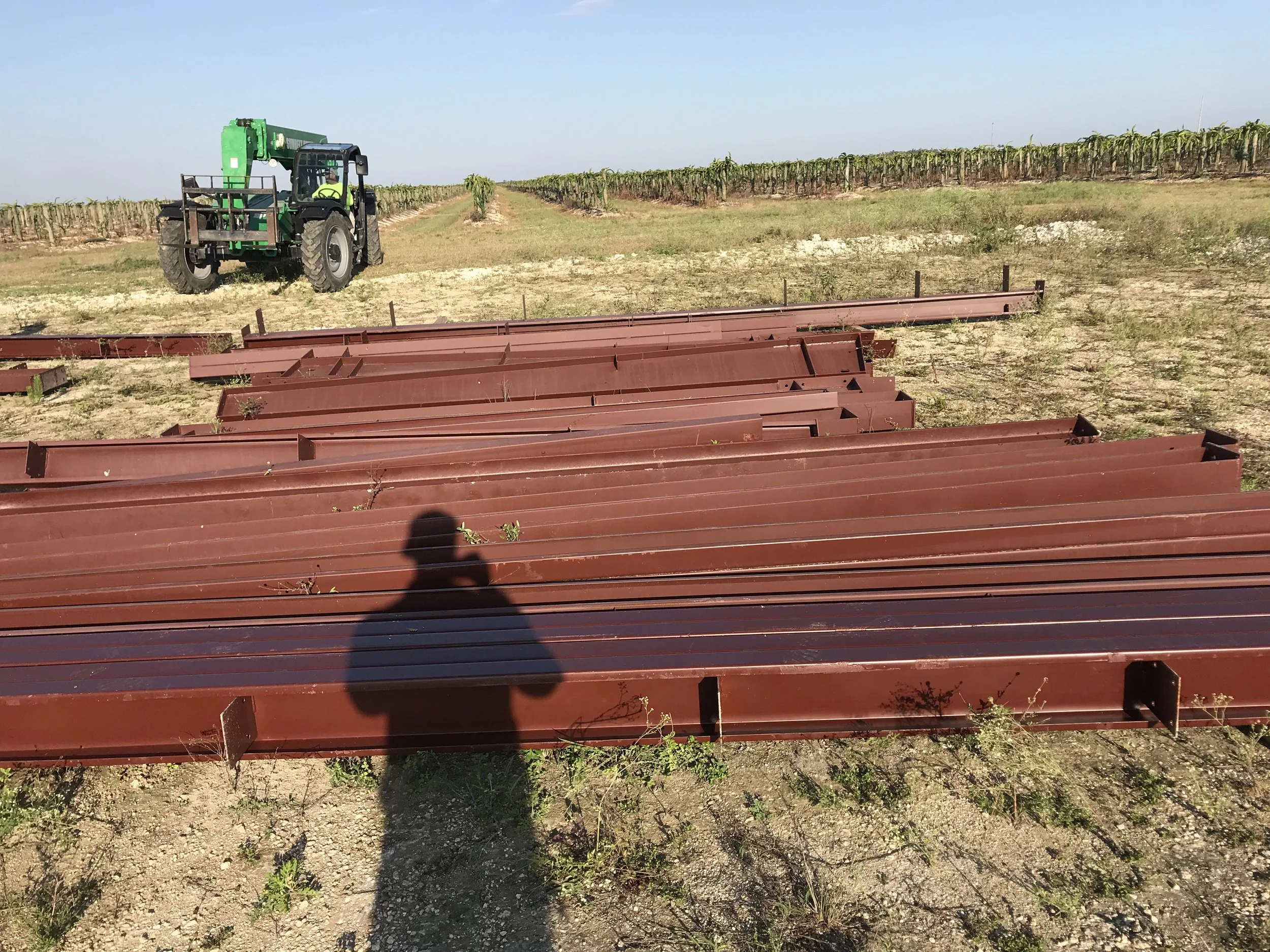 Steel beams laid out on dirt field with a tractor in distance, under clear blue sky.