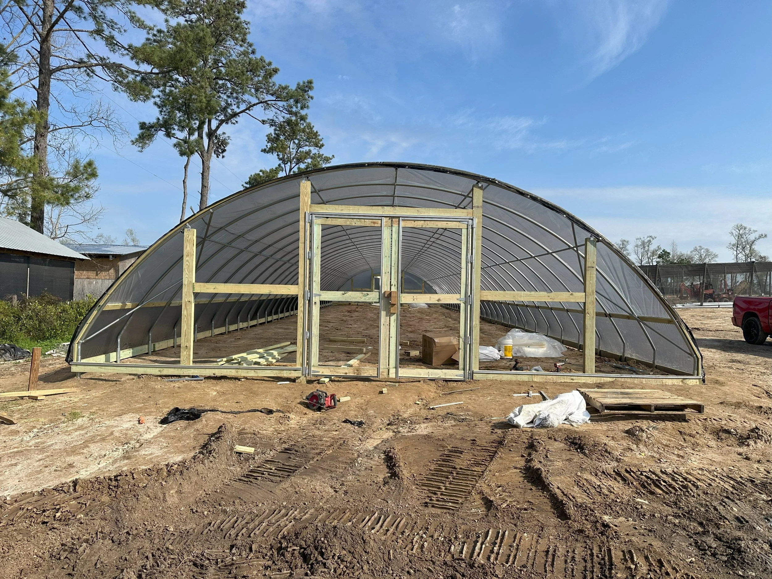 Construction of a greenhouse with a curved metal frame and transparent panels, on a dirt ground with tools and building materials scattered around, under a clear blue sky with trees in the background.