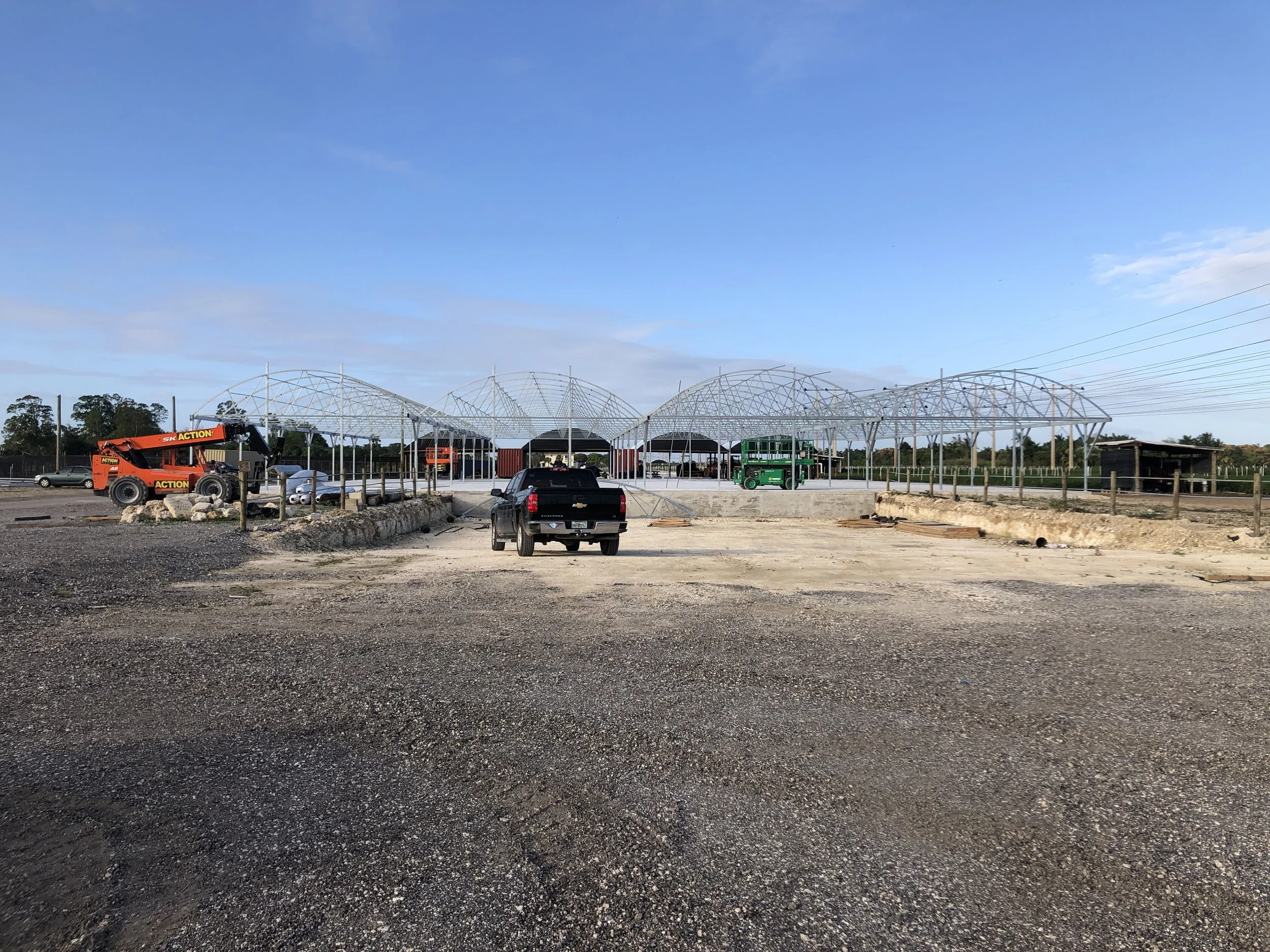 Construction site with a partially assembled large meta Green house structure, vehicles including a black pickup truck, green tractor, and orange machinery on dirt ground under a blue sky with clouds.