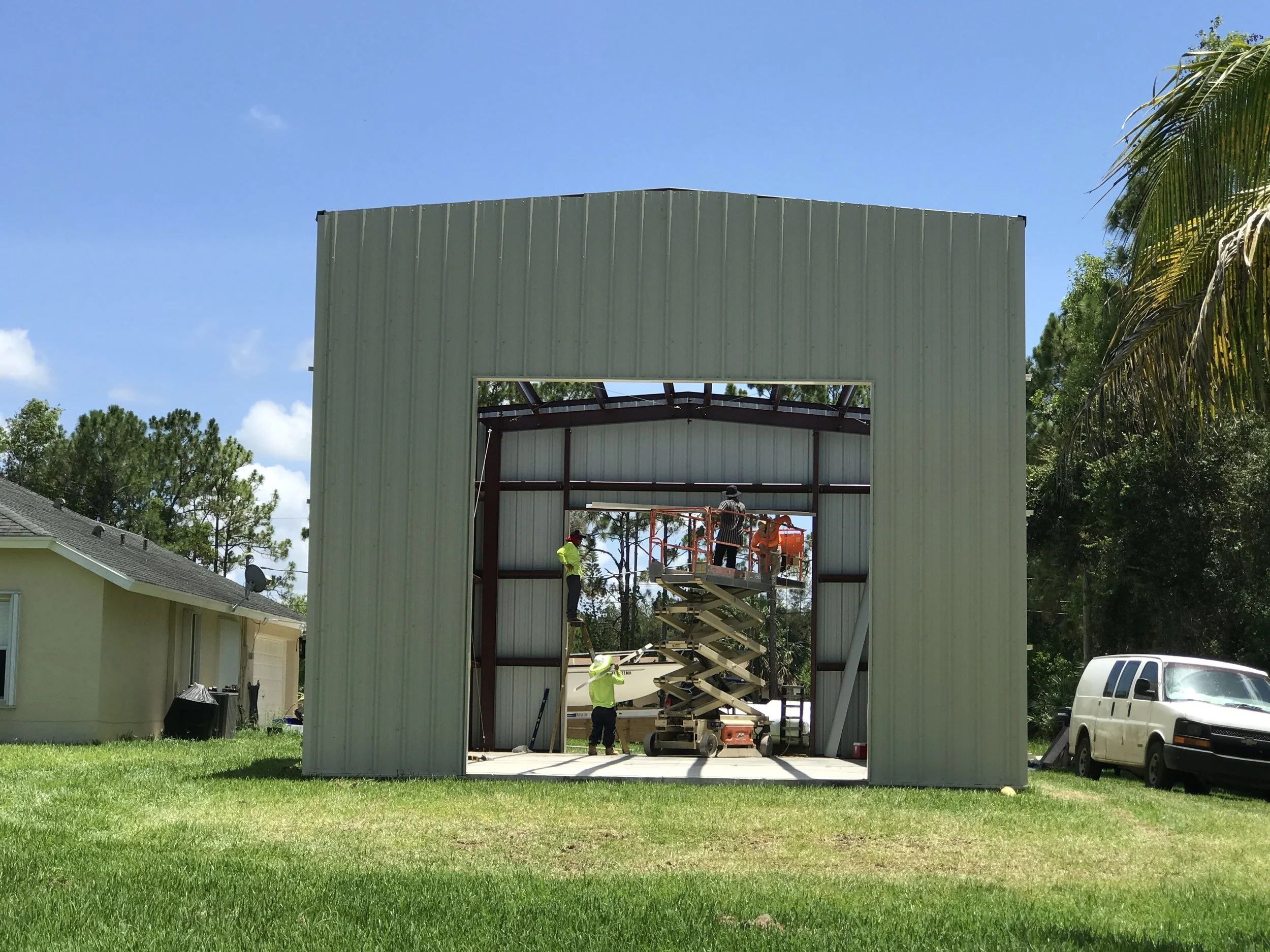 Workers constructing a metal building inside a green yard with trees and neighbors' houses nearby.