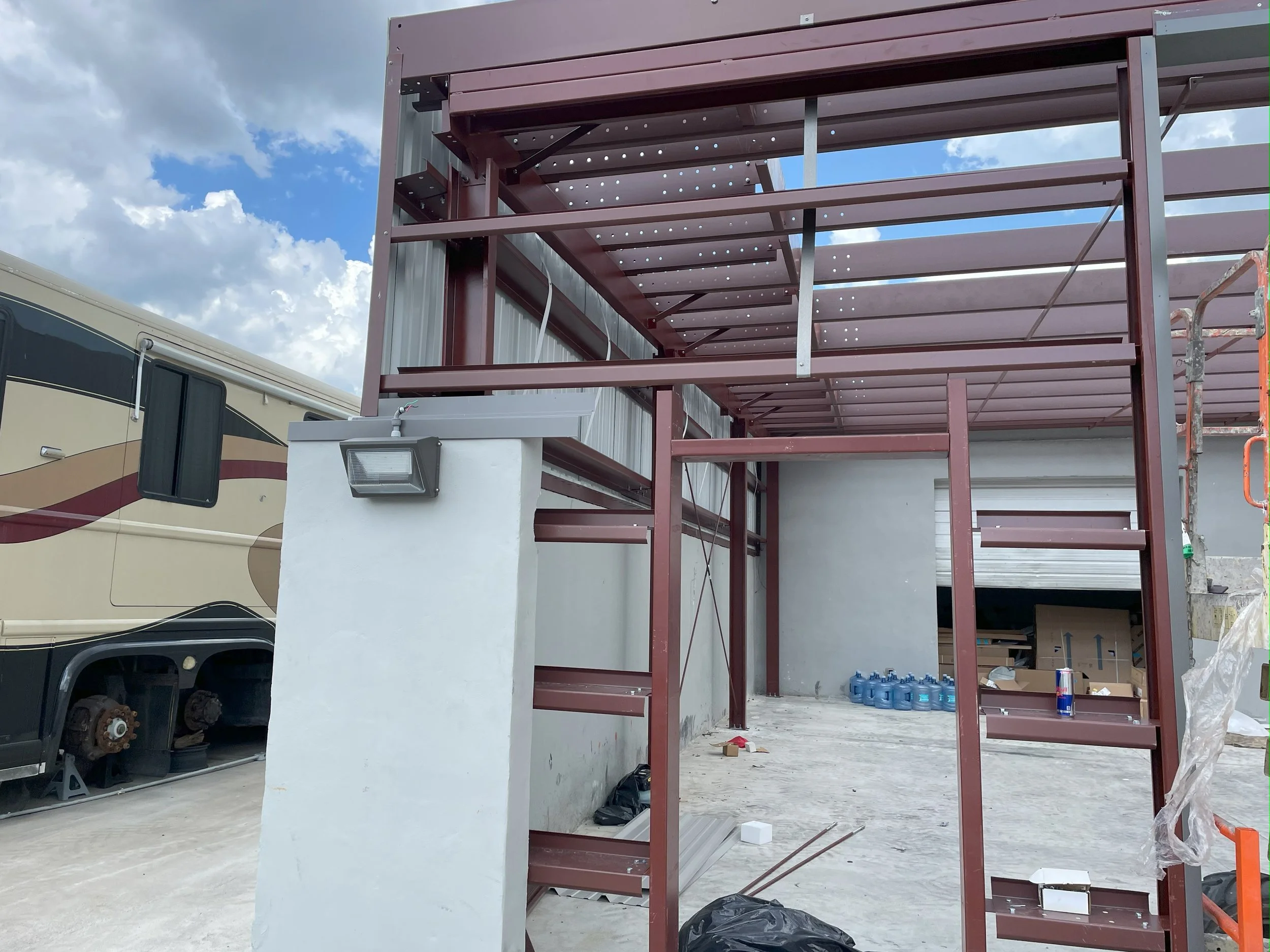Construction site with metal framing for a building, a white wall with a light fixture, and a row of water bottles inside. A recreational vehicle is parked nearby.