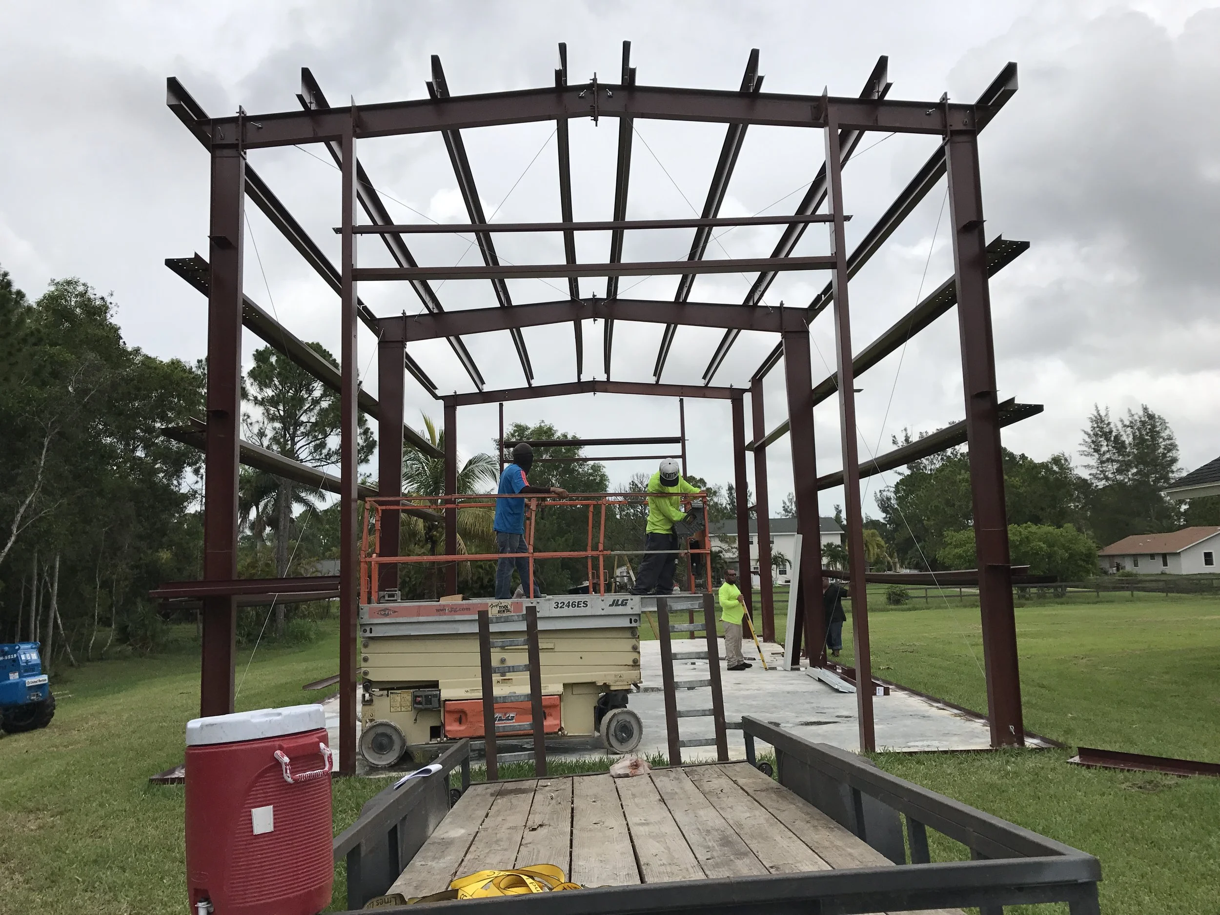 Construction workers assembling a steel frame on a building site, with some workers on a lift, in a grassy area with houses in the background.