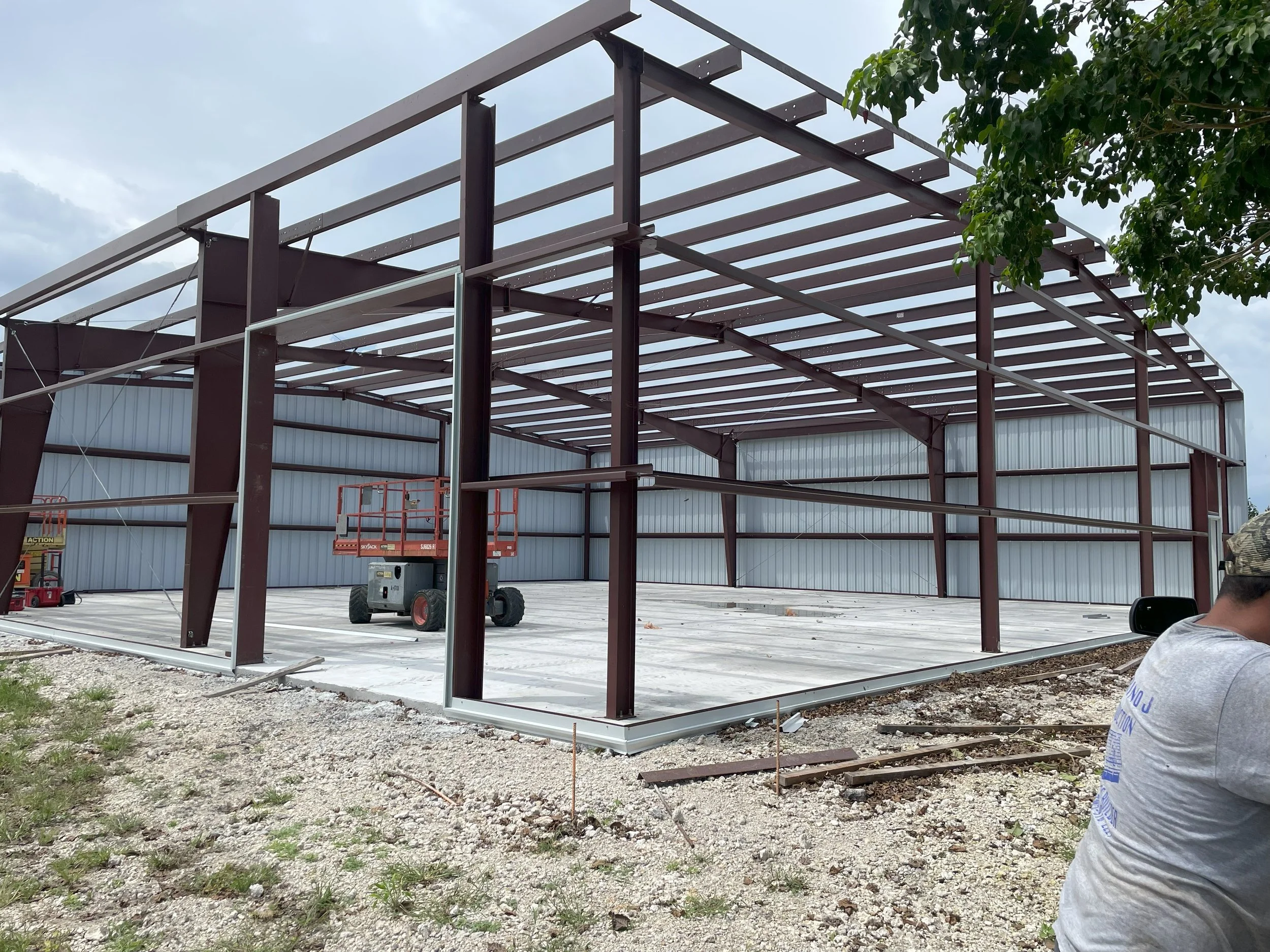 Steel framework of a building under construction with a partially installed metal roof and walls. Construction equipment and a person in a gray shirt and camouflage hat are visible.