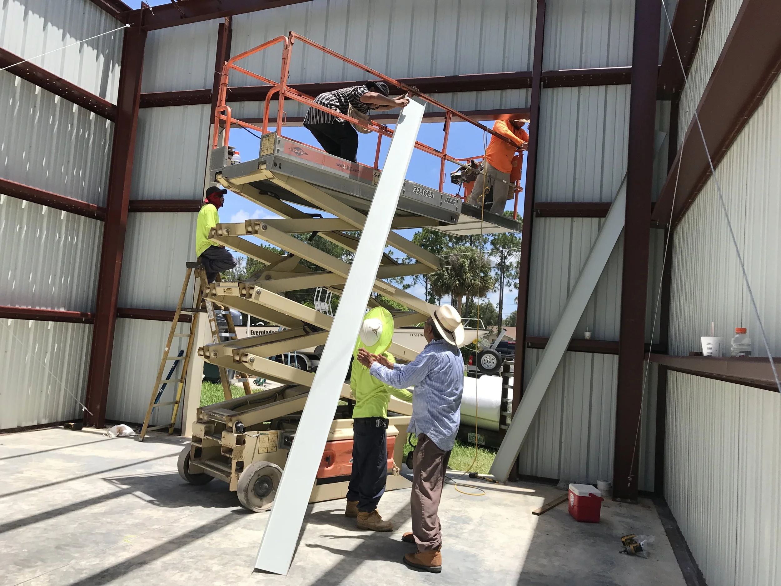 Construction workers using a scissor lift inside a metal building frame, with two workers on the lift and two on the ground, and a person in a hat clapping.