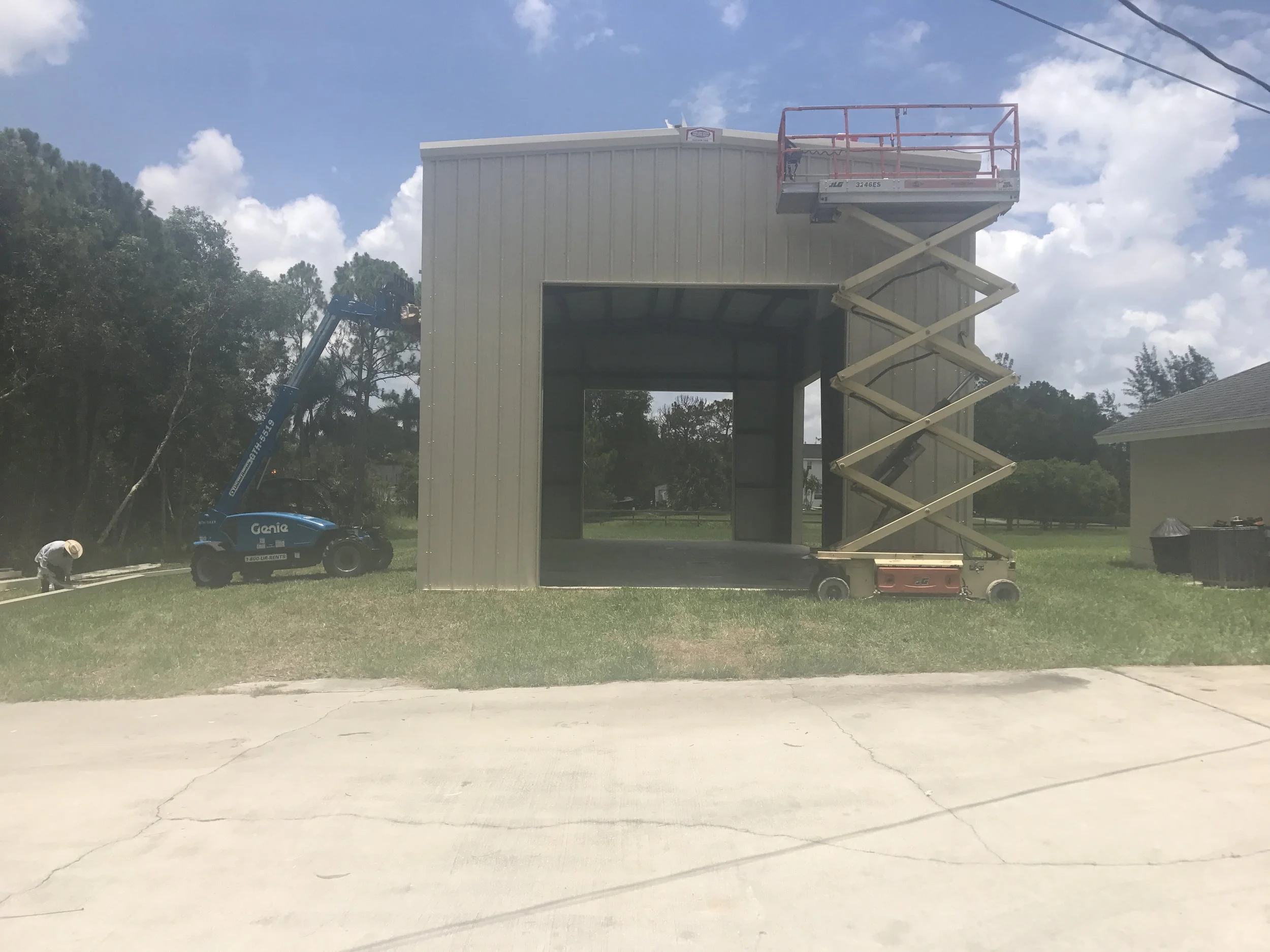 Construction site with a metal shed, a blue Genie lift with a worker, and a scissor lift on the ground, under a partly cloudy sky.
