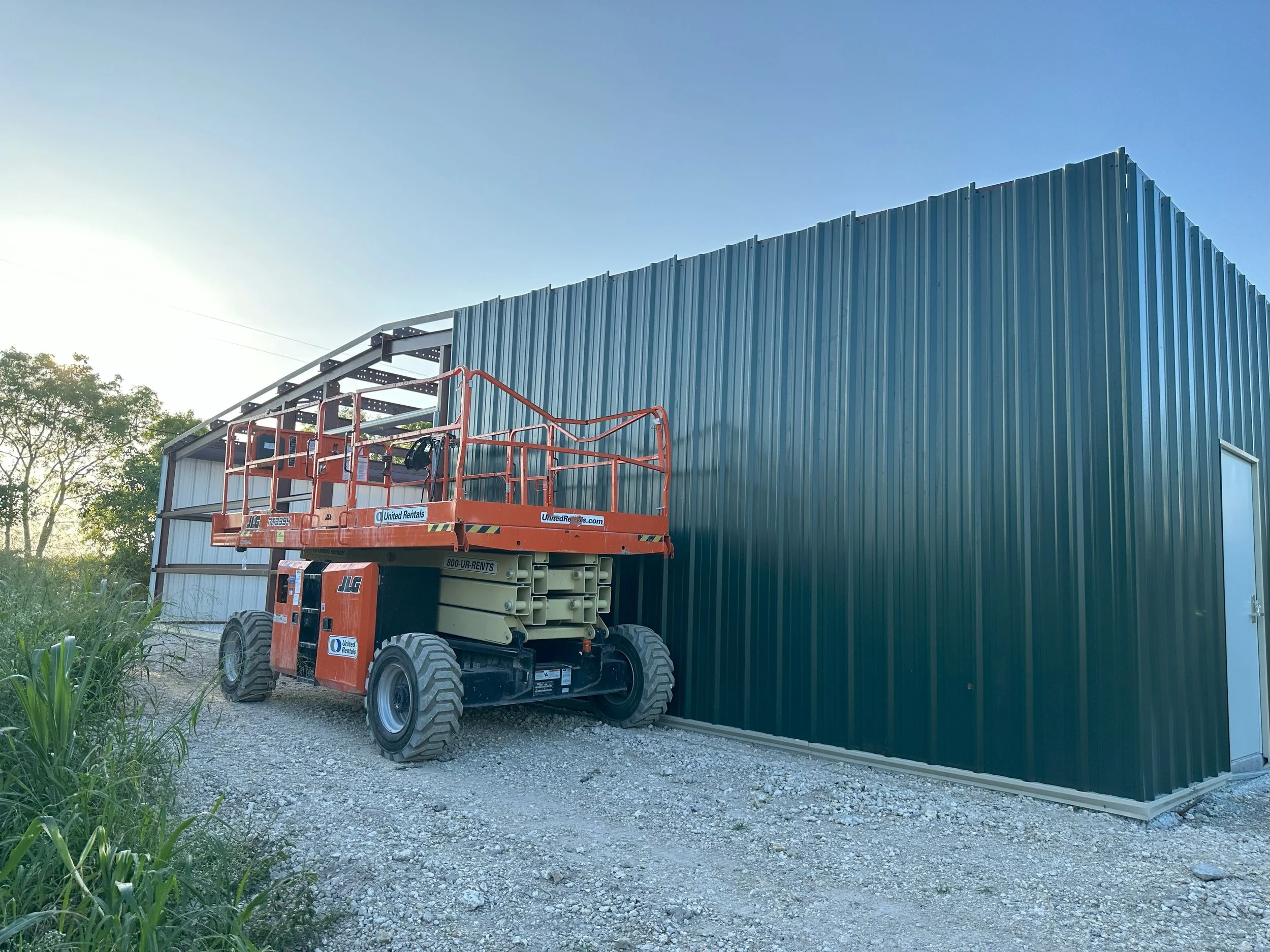 Construction site with a large green metal building and an orange scissor lift parked outside.
