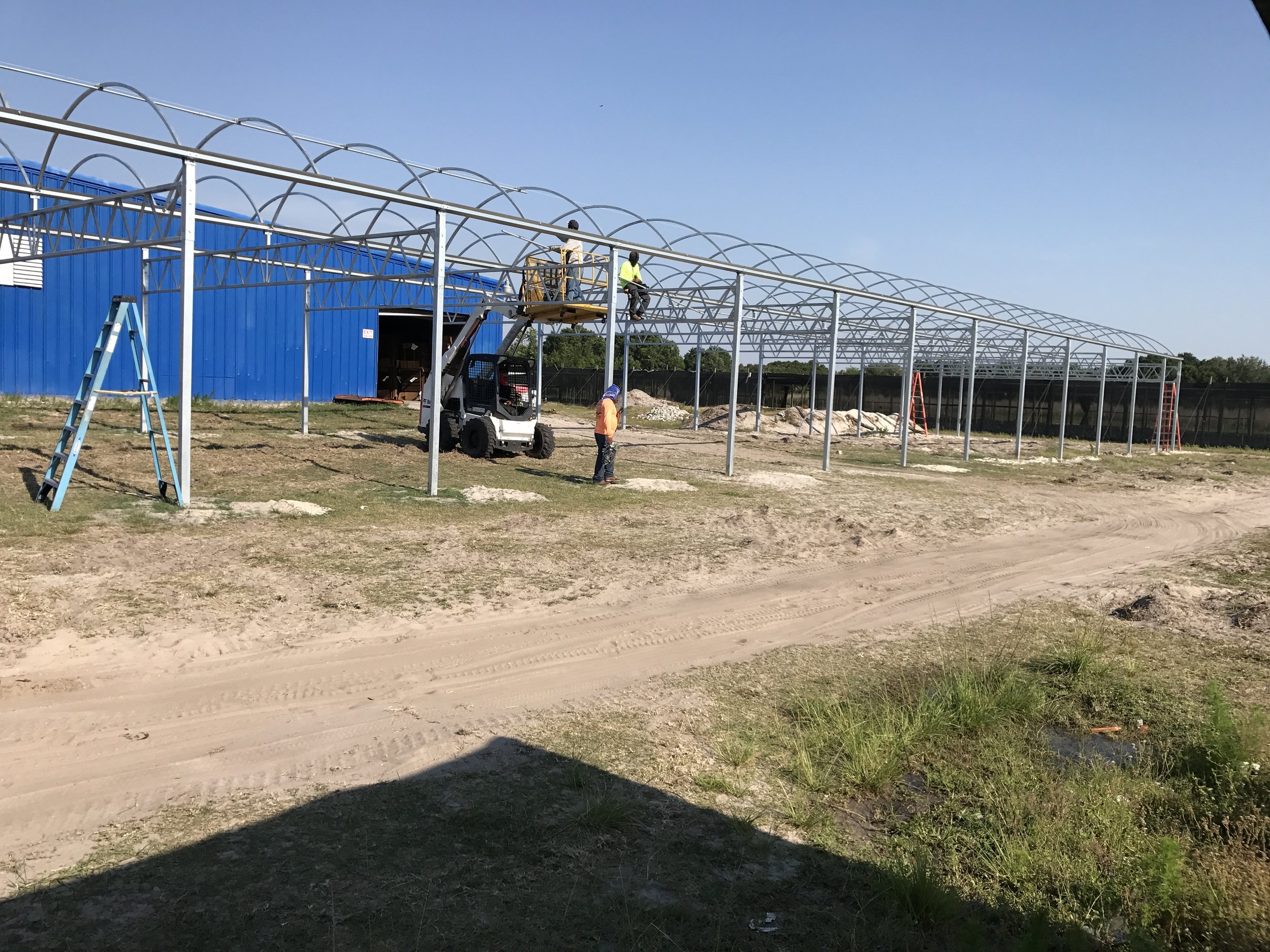 Construction workers assembling a metal framework structure outdoors on a dirt lot, with a blue building in the background.