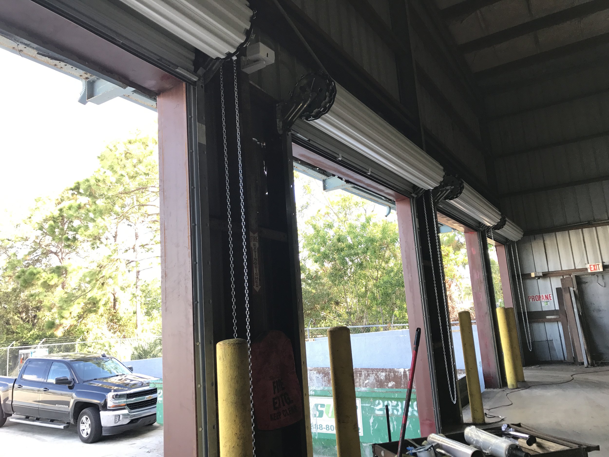 Open garage bay door with a black pickup truck outside, trees visible, and part of the indoor shop area with equipment and yellow safety bollards.