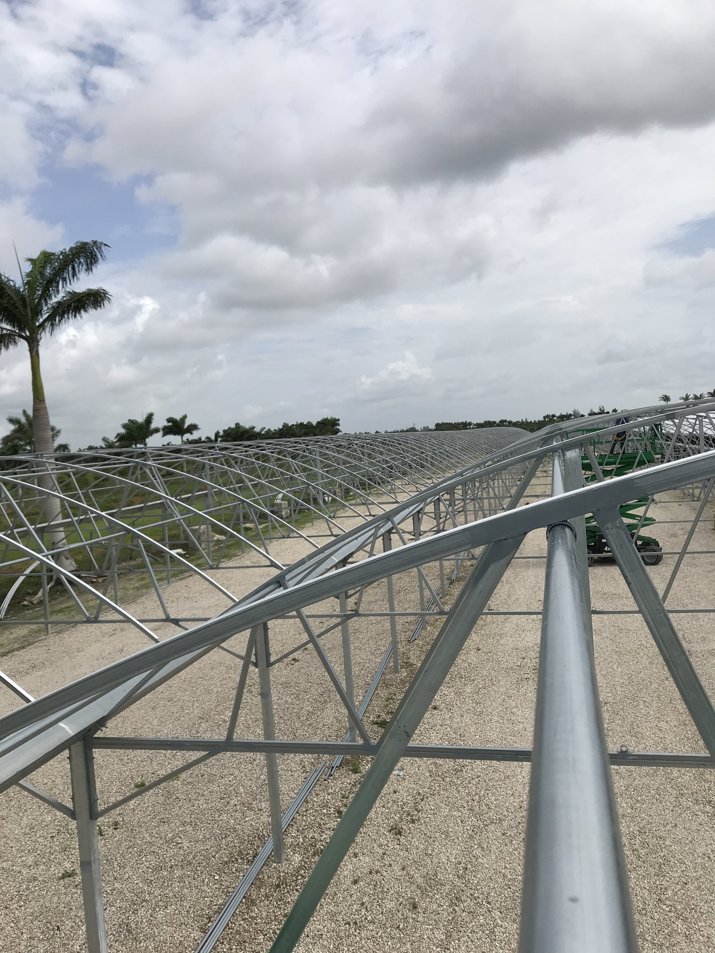 Metal framework for solar panels being installed outdoors with palm trees and cloudy sky in the background.