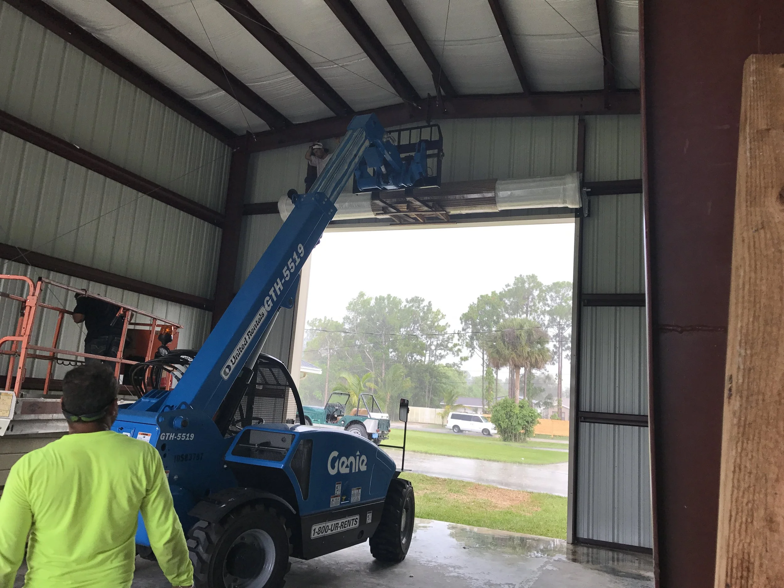 Worker in a yellow shirt operating a blue Genie GTH-5519 telehandler inside a large metal building, with the boom raised near the ceiling and a person in a helmet on a platform above.