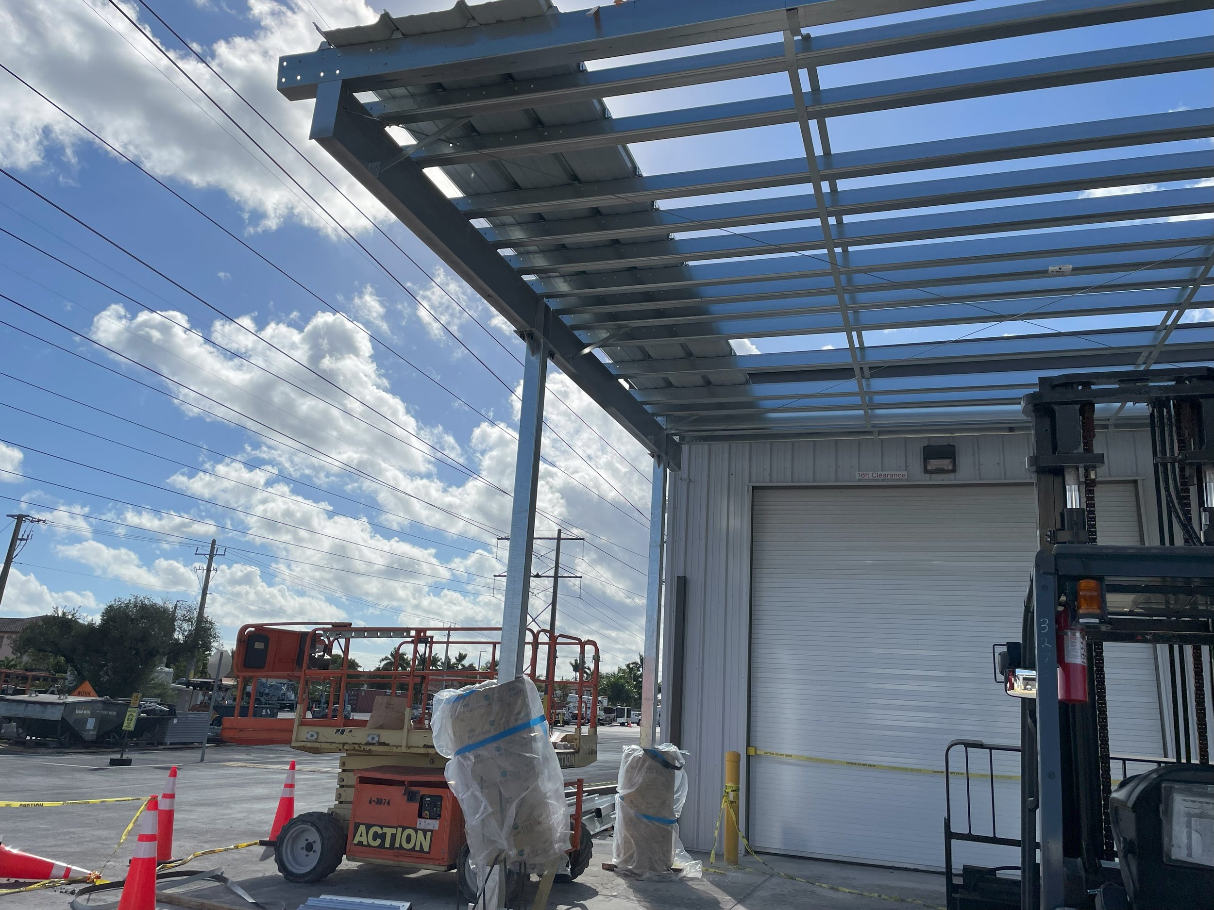 Construction site with a partially built metal awning, a forklift, a scissor lift, safety cones, and a large shutter door on a sunny day with partly cloudy skies.