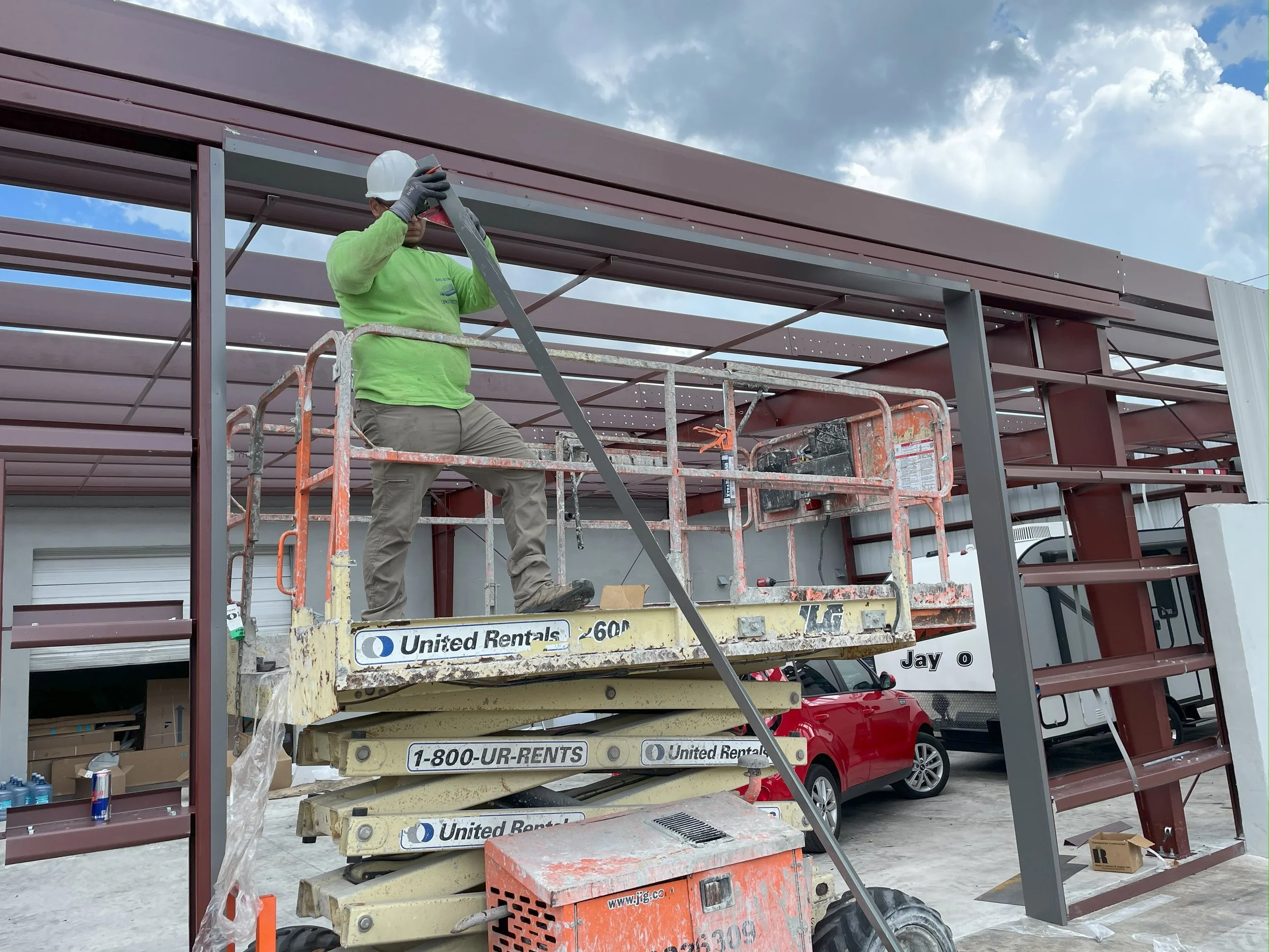 Construction worker in lime green shirt, gray pants, and white helmet standing on a scissor lift, installing steel beams on a building under construction, with a red car and a white RV parked in the background under a partly cloudy sky.