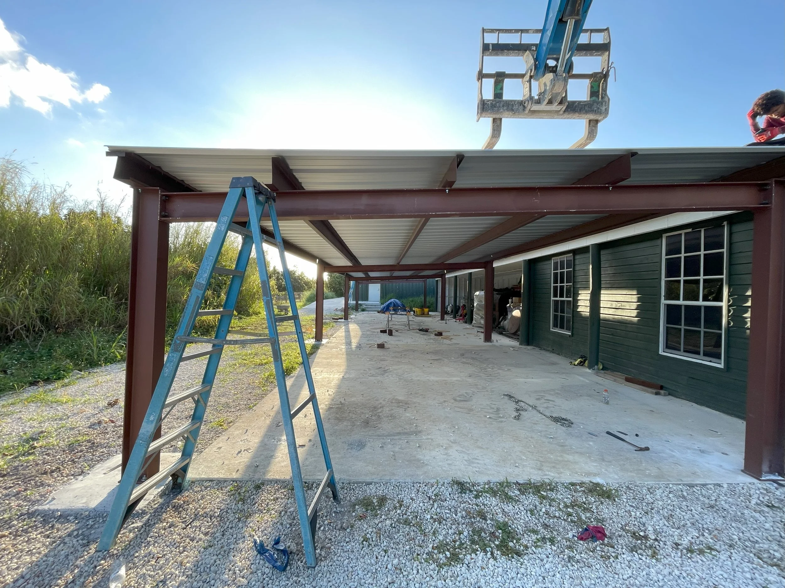 Construction site with a carport being built, showing steel beams and a corrugated metal roof, with a blue ladder in the foreground and construction tools scattered on the ground, adjacent to a green house with white-framed windows.