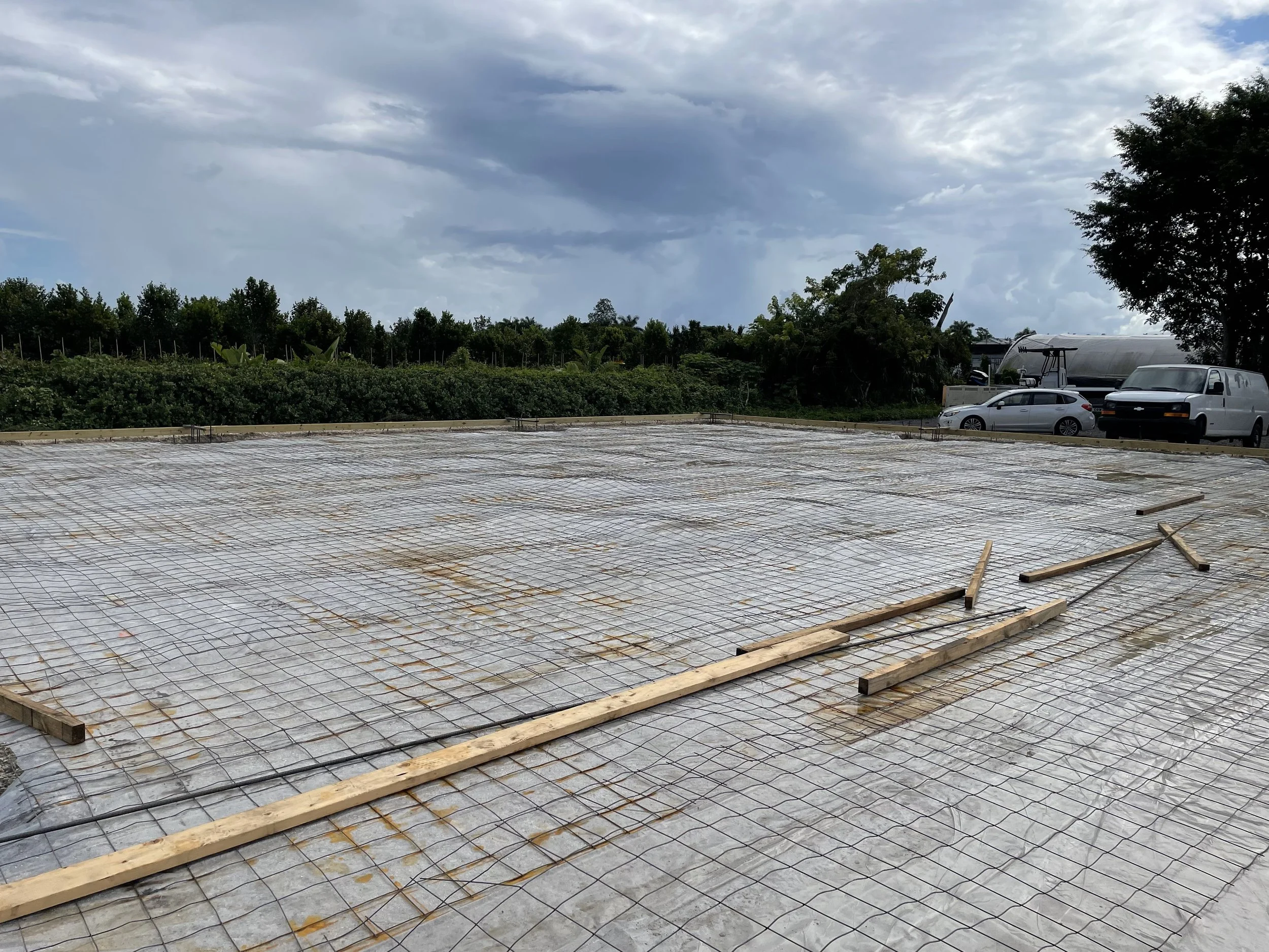 Construction site with rebar reinforcement laid out on the ground, covered with plastic sheeting, with cars and trees in the background under cloudy skies.