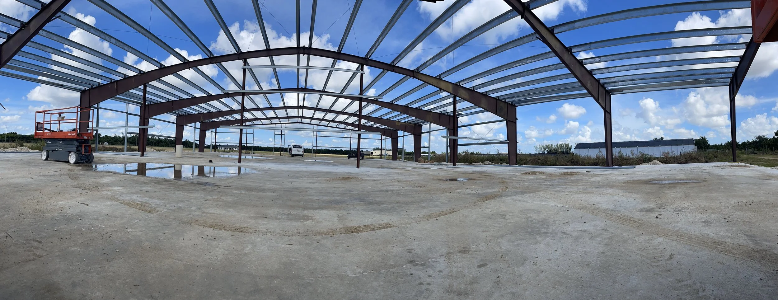 Construction site with a large steel frame structure and a clear blue sky with scattered clouds, concrete floor with puddles, and construction equipment.