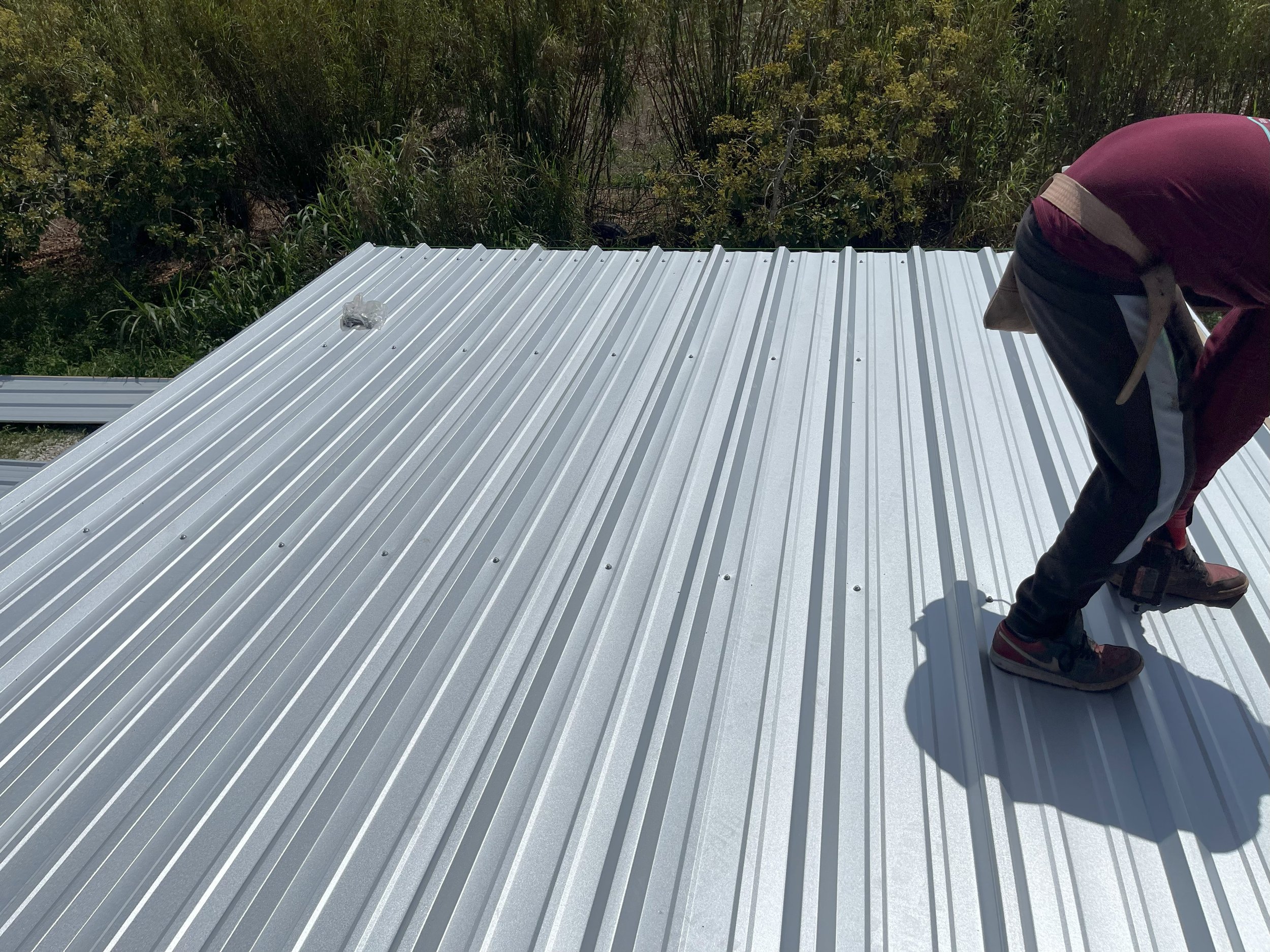 Person installing or working on a silver, ridged metal roof outdoors, with trees and bushes in the background.