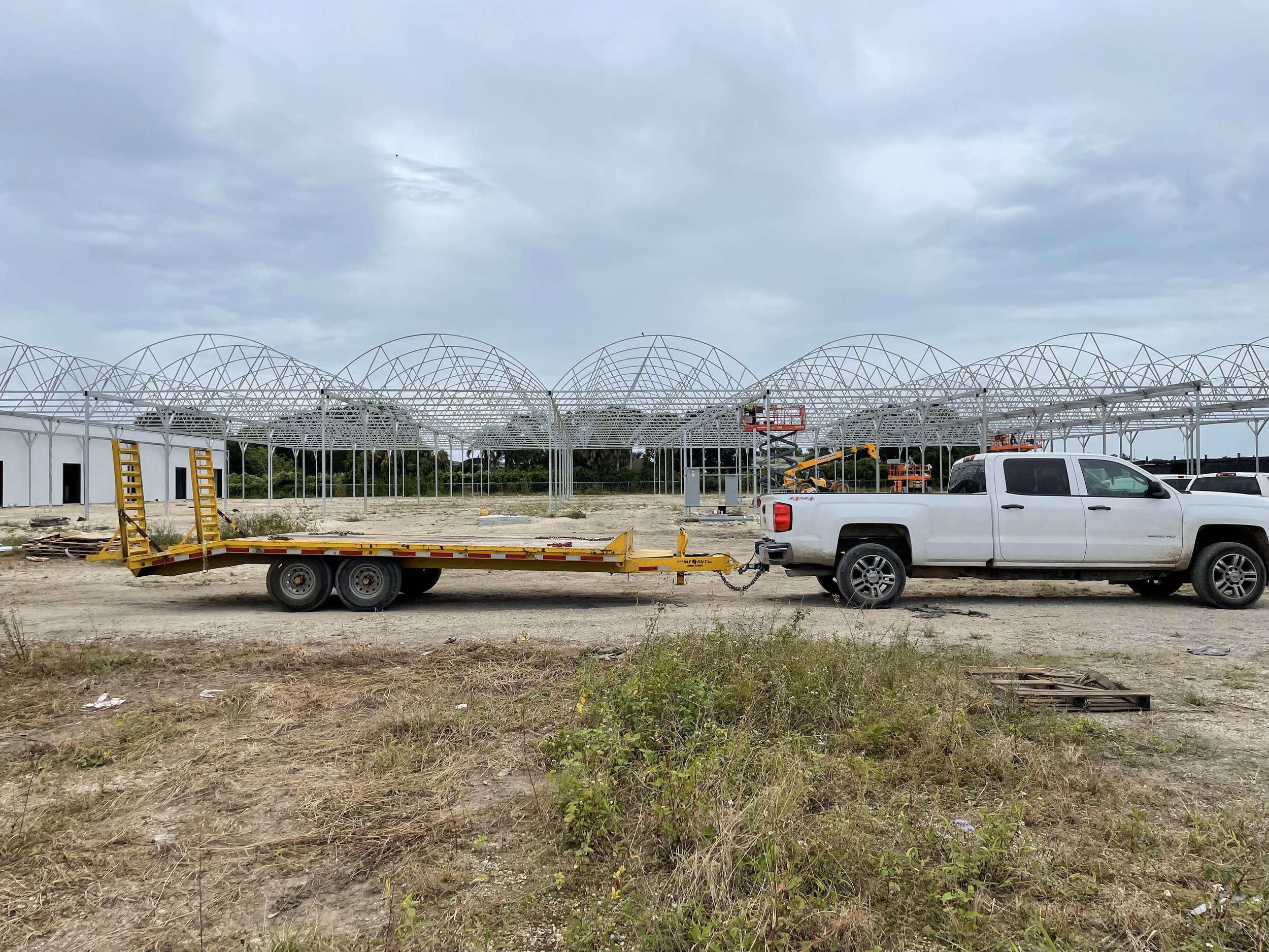 A white pickup truck with a green utility bed dragging a flatbed trailer at a construction site, with a large metal frame structure and construction equipment in the background under cloudy sky.