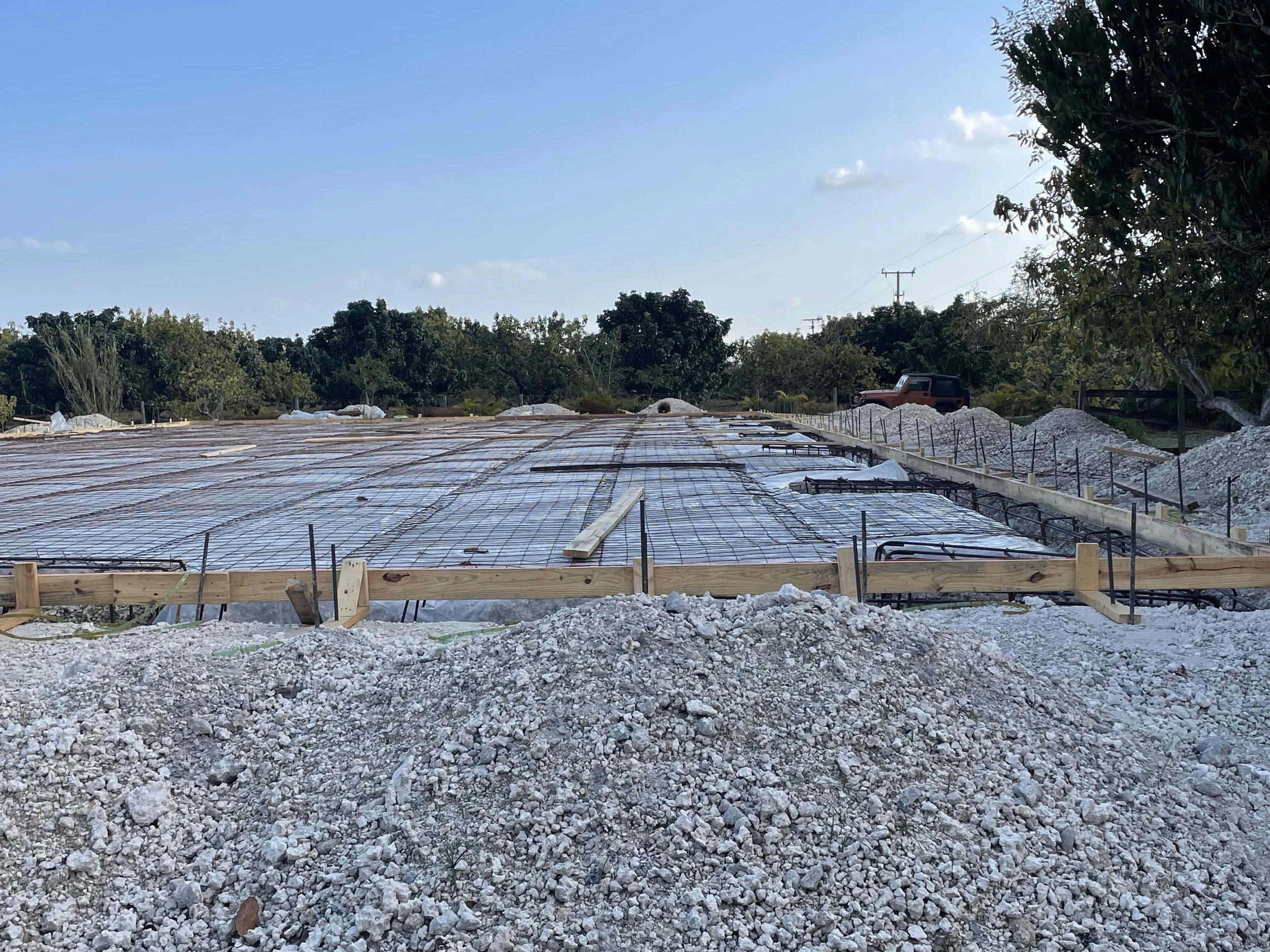Construction site with a concrete foundation, rebar, and wooden formwork, surrounded by mounds of gravel and trees in the background.