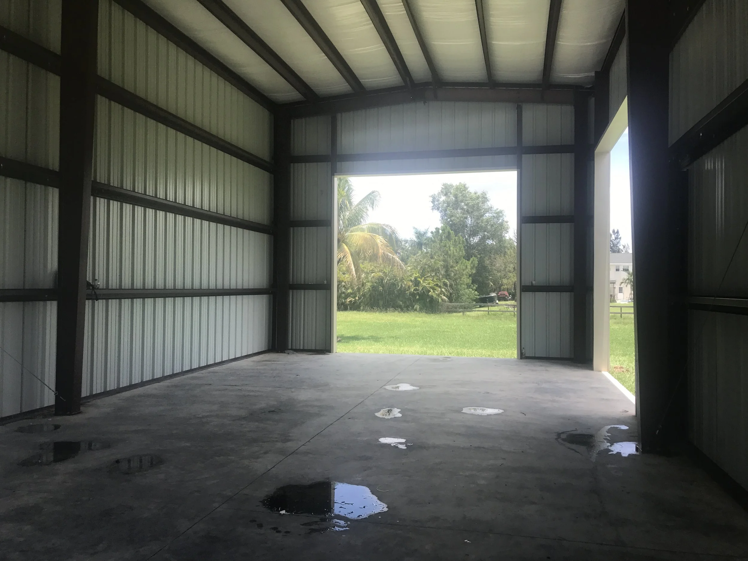 Interior of an empty metal storage or garage building with concrete floor, open garage door showing green trees and grass outside, and some water puddles on the floor.