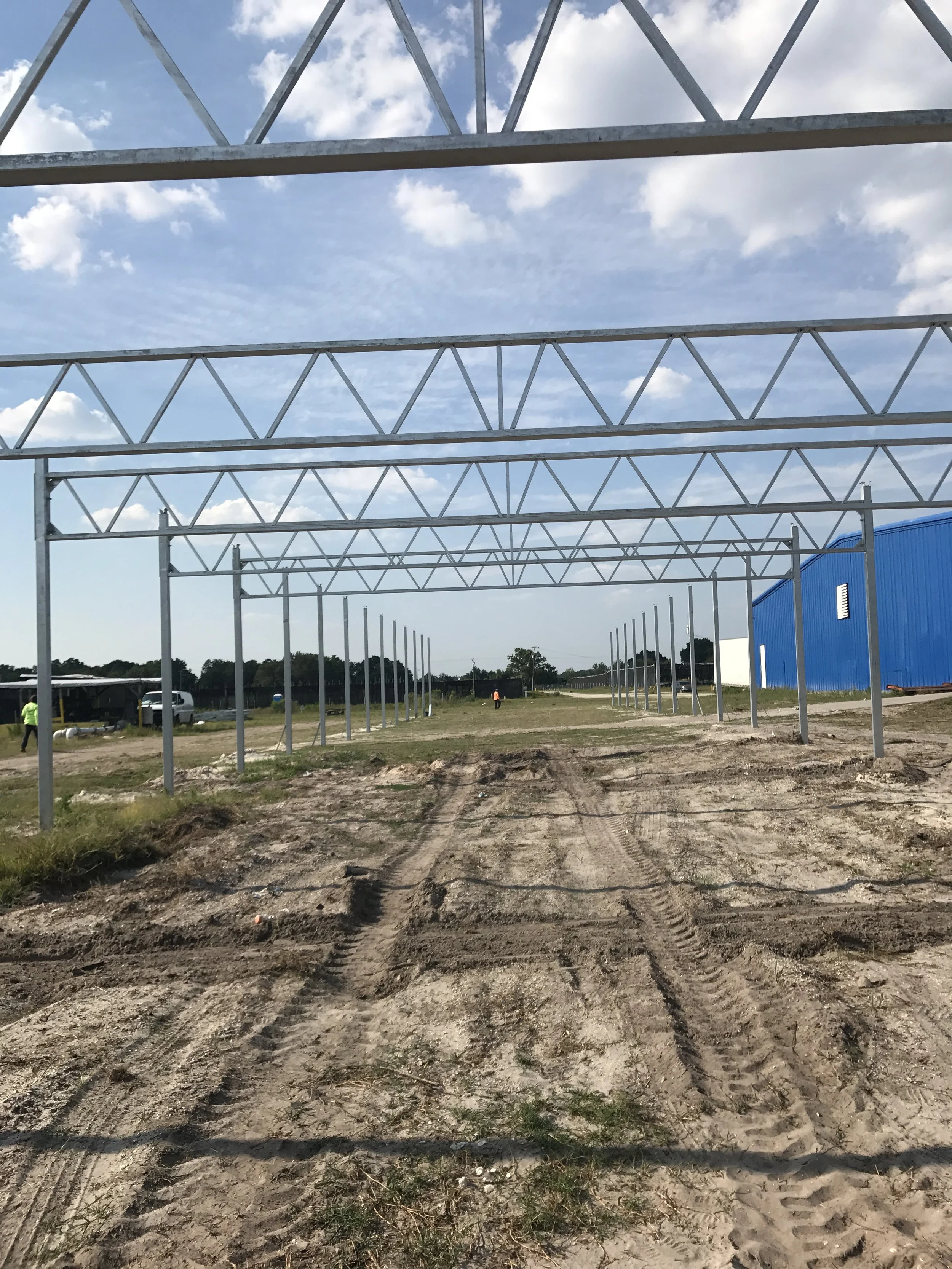Construction site with metal framework for a building, dirt ground with tire tracks, blue sky with scattered clouds.