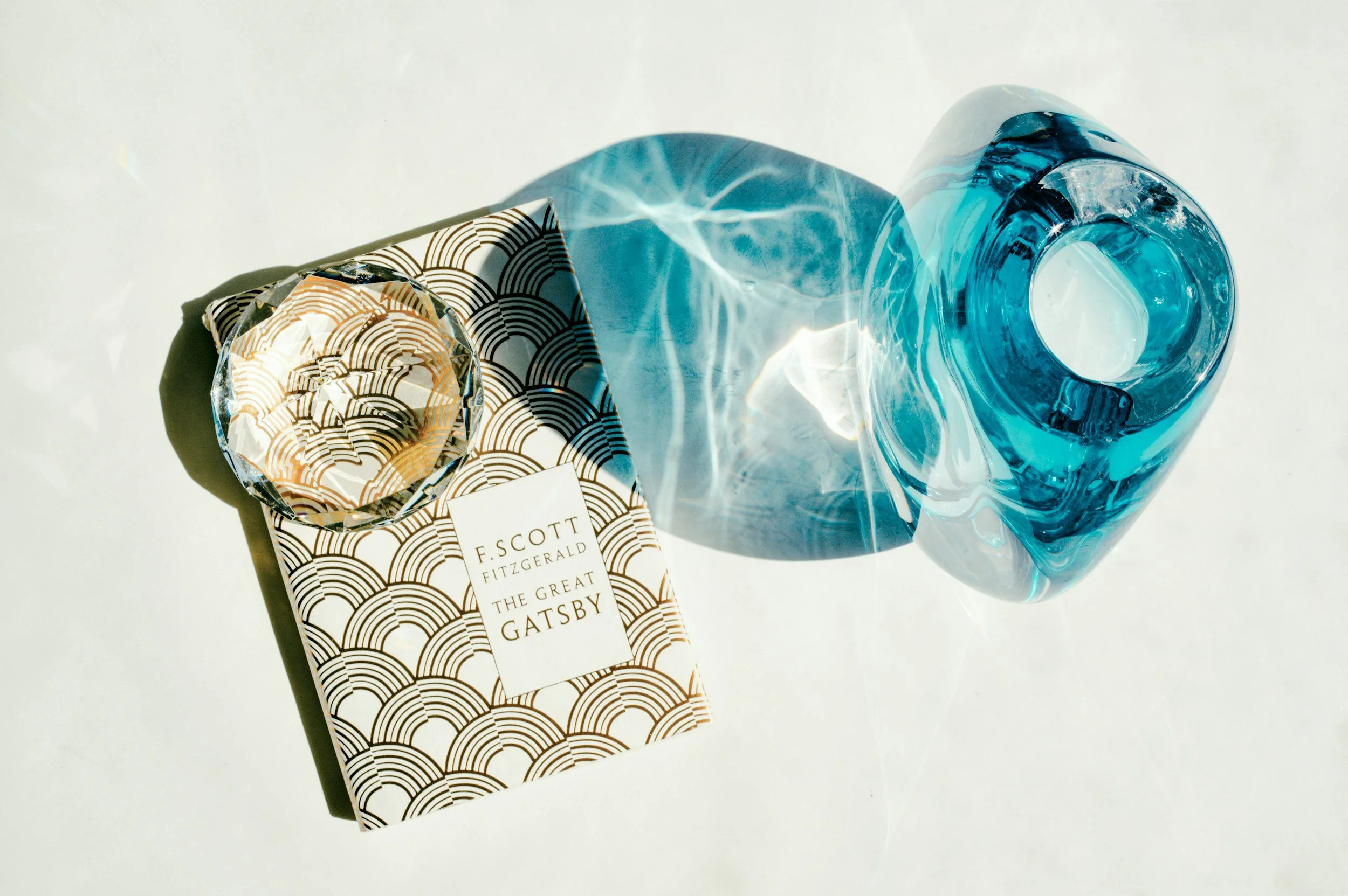 A copy of 'The Great Gatsby' book with a patterned cover and a decorative glass of blue water on a white surface.