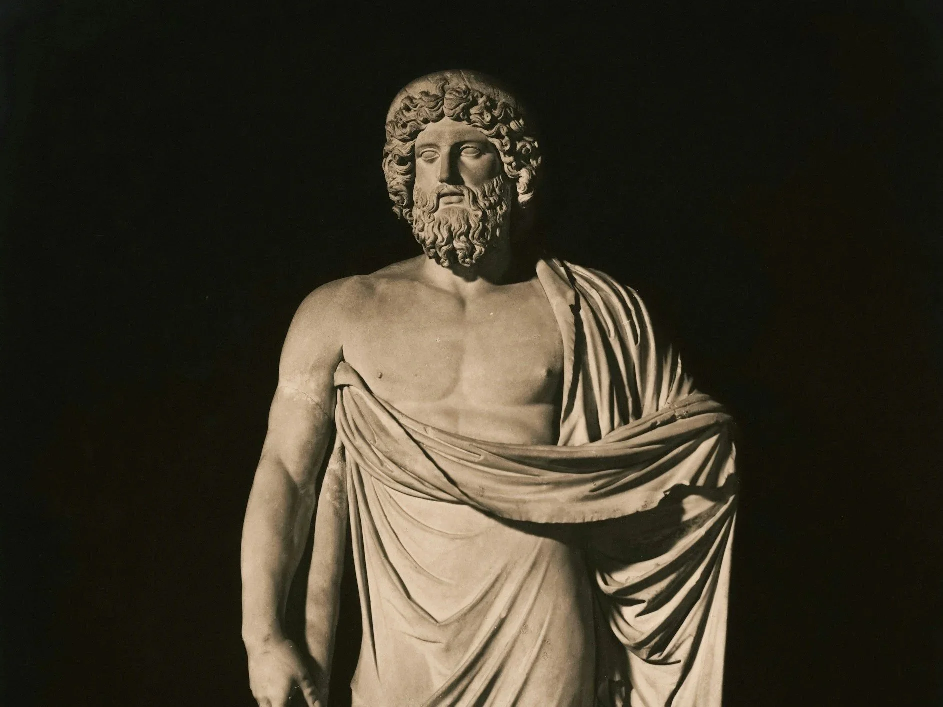 A black and white photograph of a classical sculpture of a bearded man with curly hair, partially draped in cloth, set against a dark background.