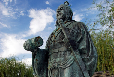 Statue of a historical figure holding a scroll and a sword, outdoors with trees and cloudy sky in the background.