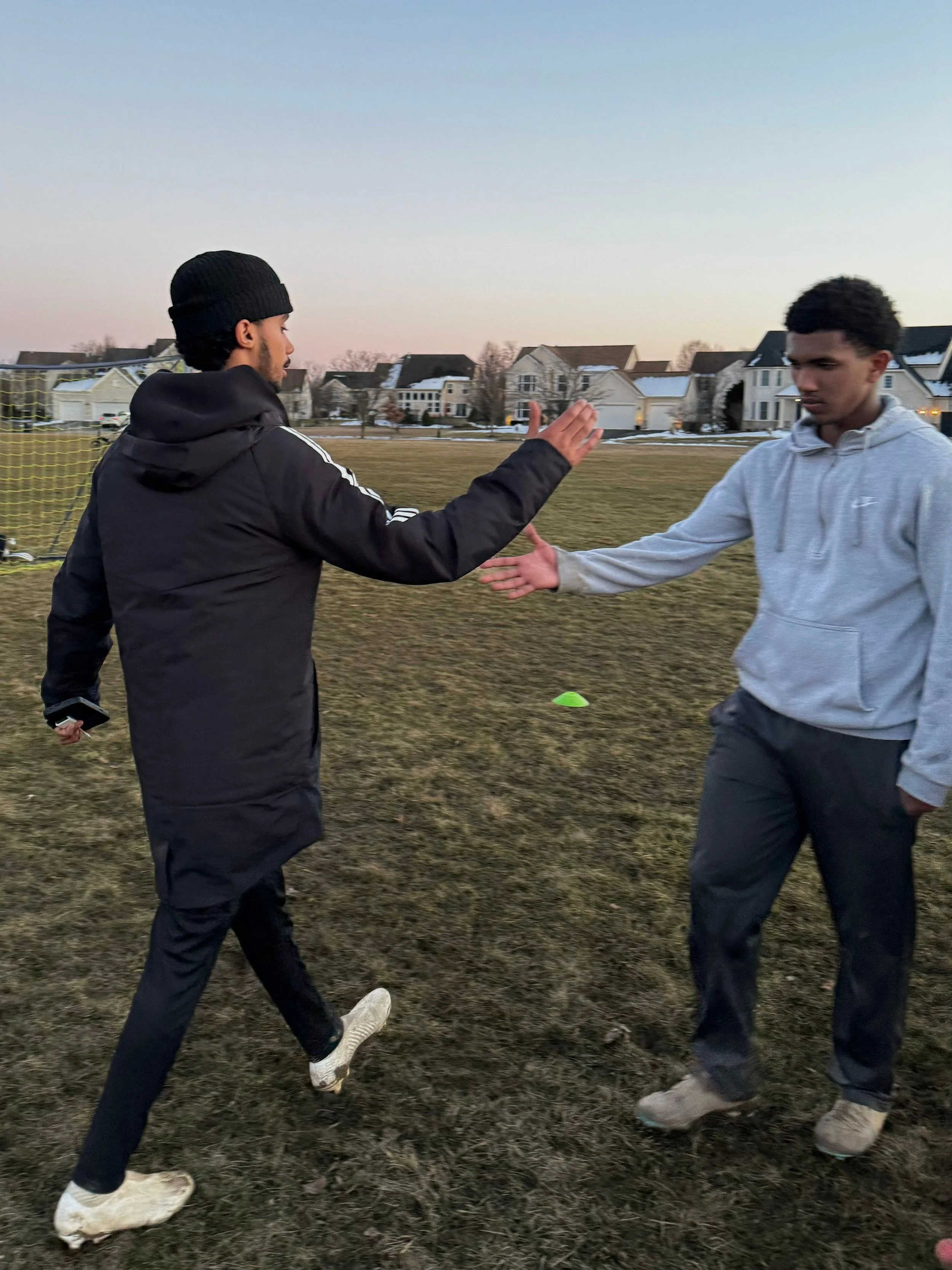 Two young men in athletic clothing greeting each other with a handshake on a grassy field during sunset