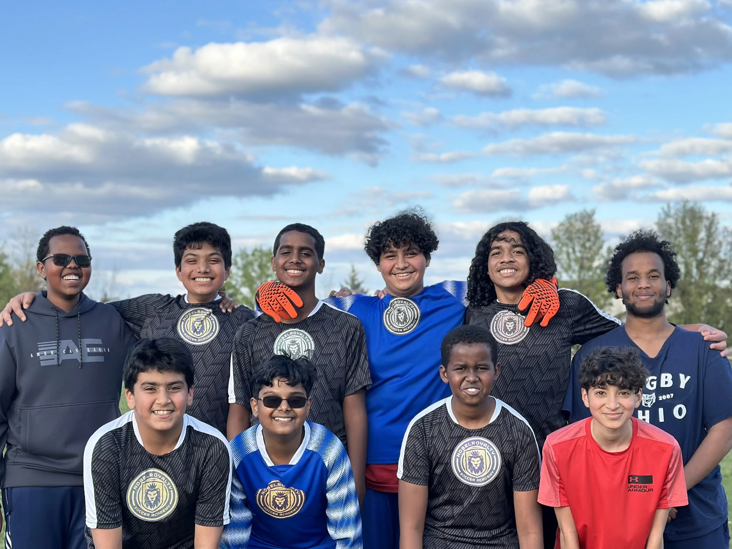 Group of young boys and a man standing outdoors on a soccer field, smiling for a photo, with a partly cloudy sky in the background.