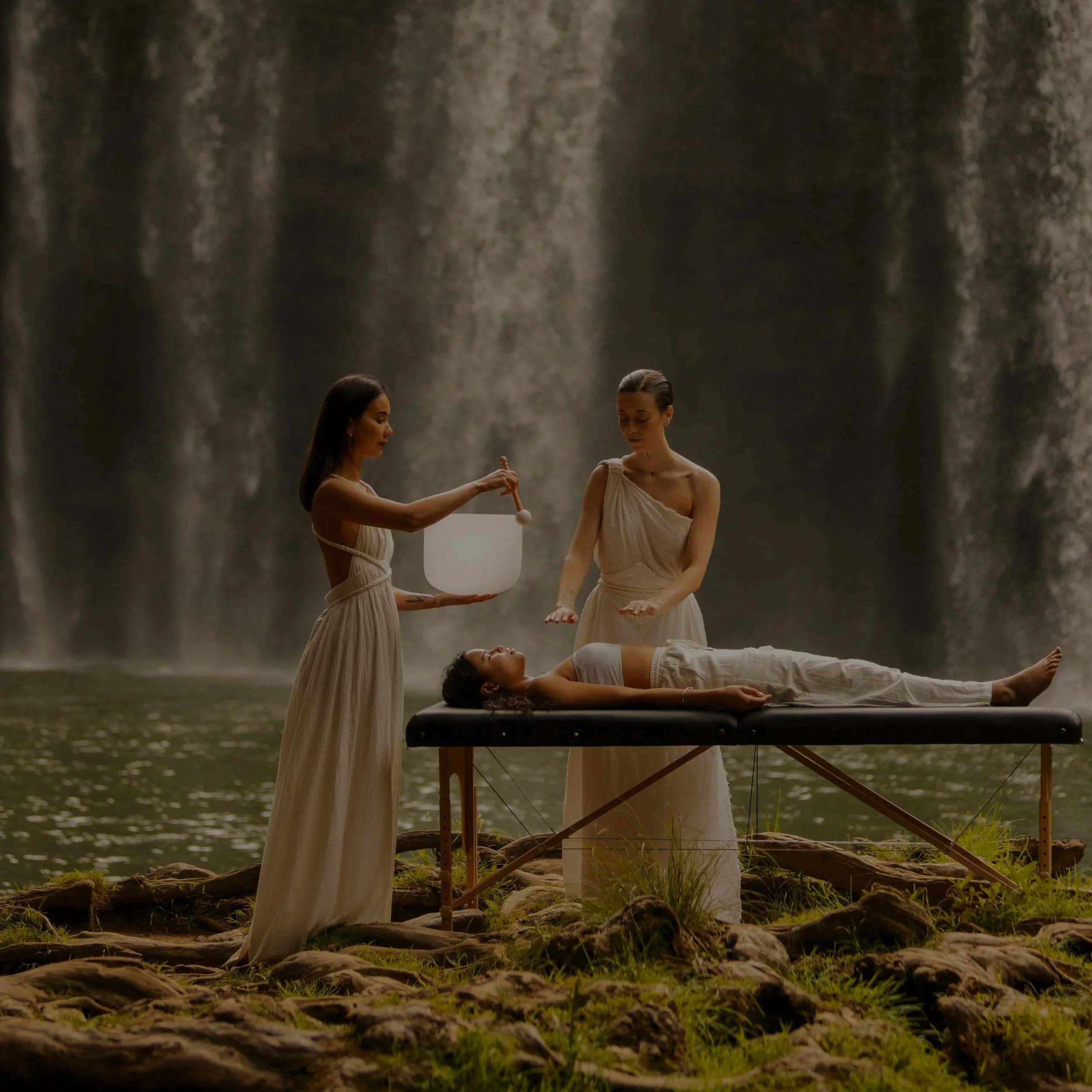 Two women standing in a dimly lit room with a black table between them. One woman on the left holds a burning object while the other on the right holds a small jar. On the table are two lit candles, a vase with flowers, and various objects, creating a warm and intimate atmosphere.