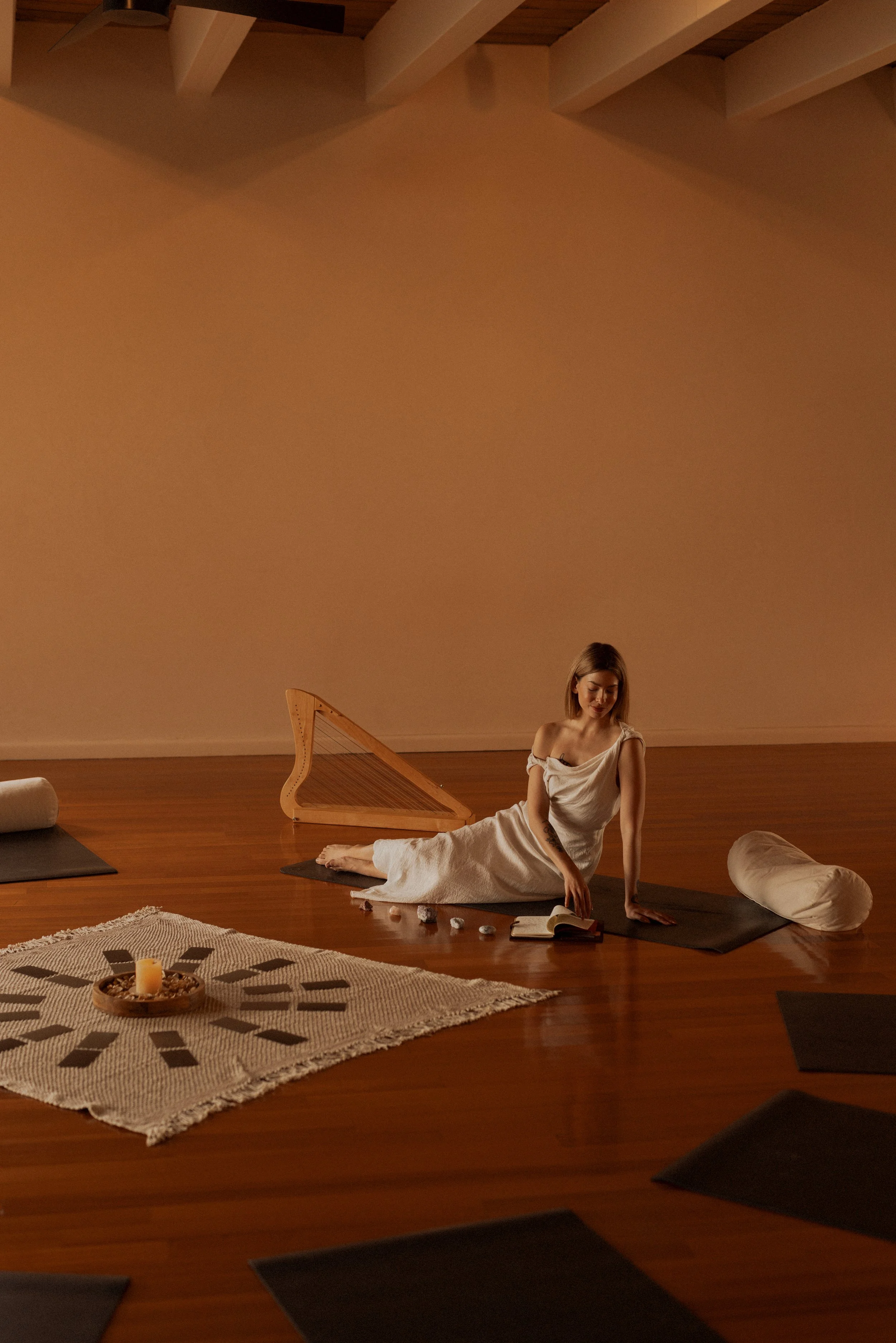 A woman in a white dress sitting on a yoga mat in a dimly lit room with wooden floors, a harp in the background, and candles and crystals on the floor in front of her.