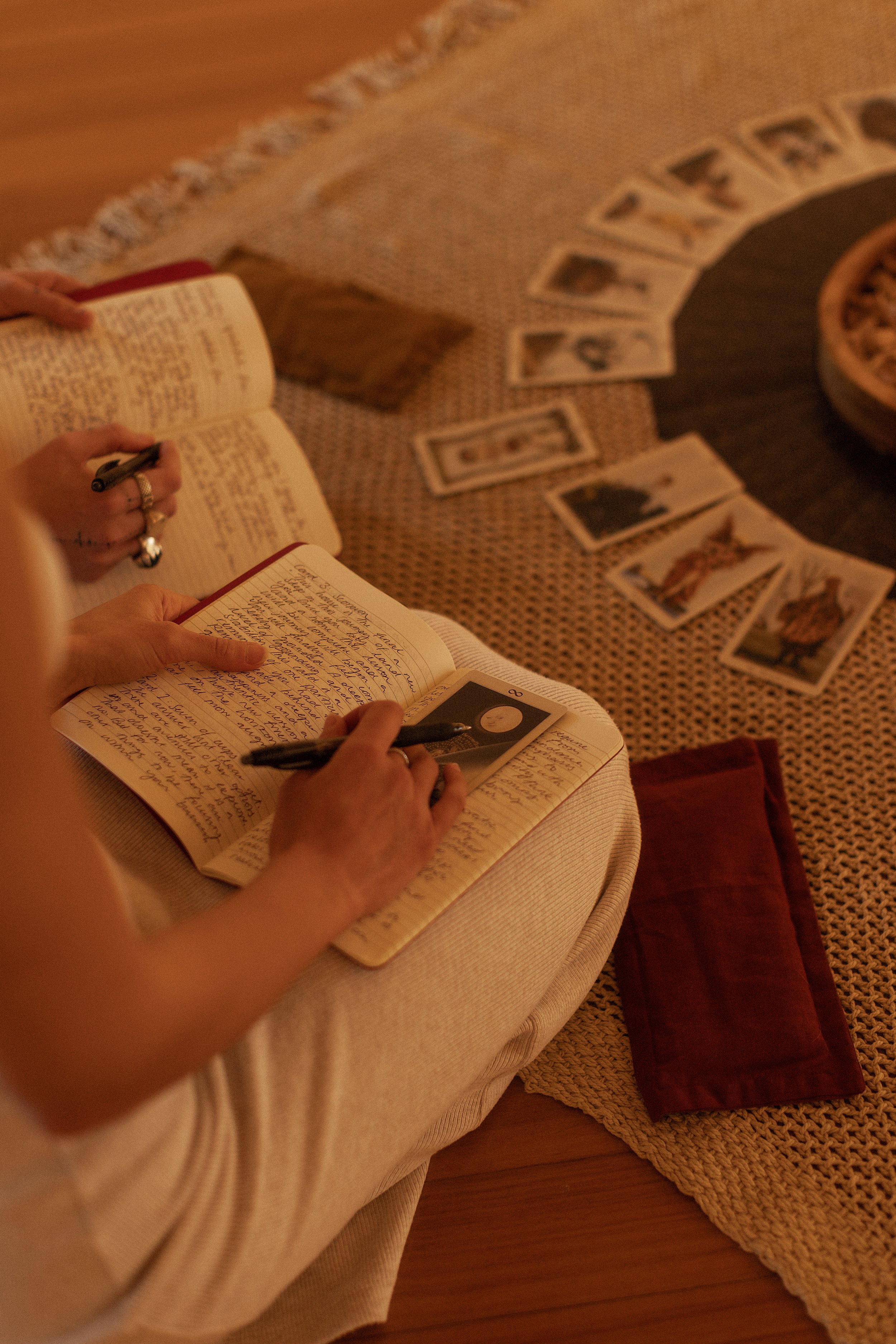Person writing in a notebook with a pen, surrounded by photographs and tarot cards on a woven tablecloth.
