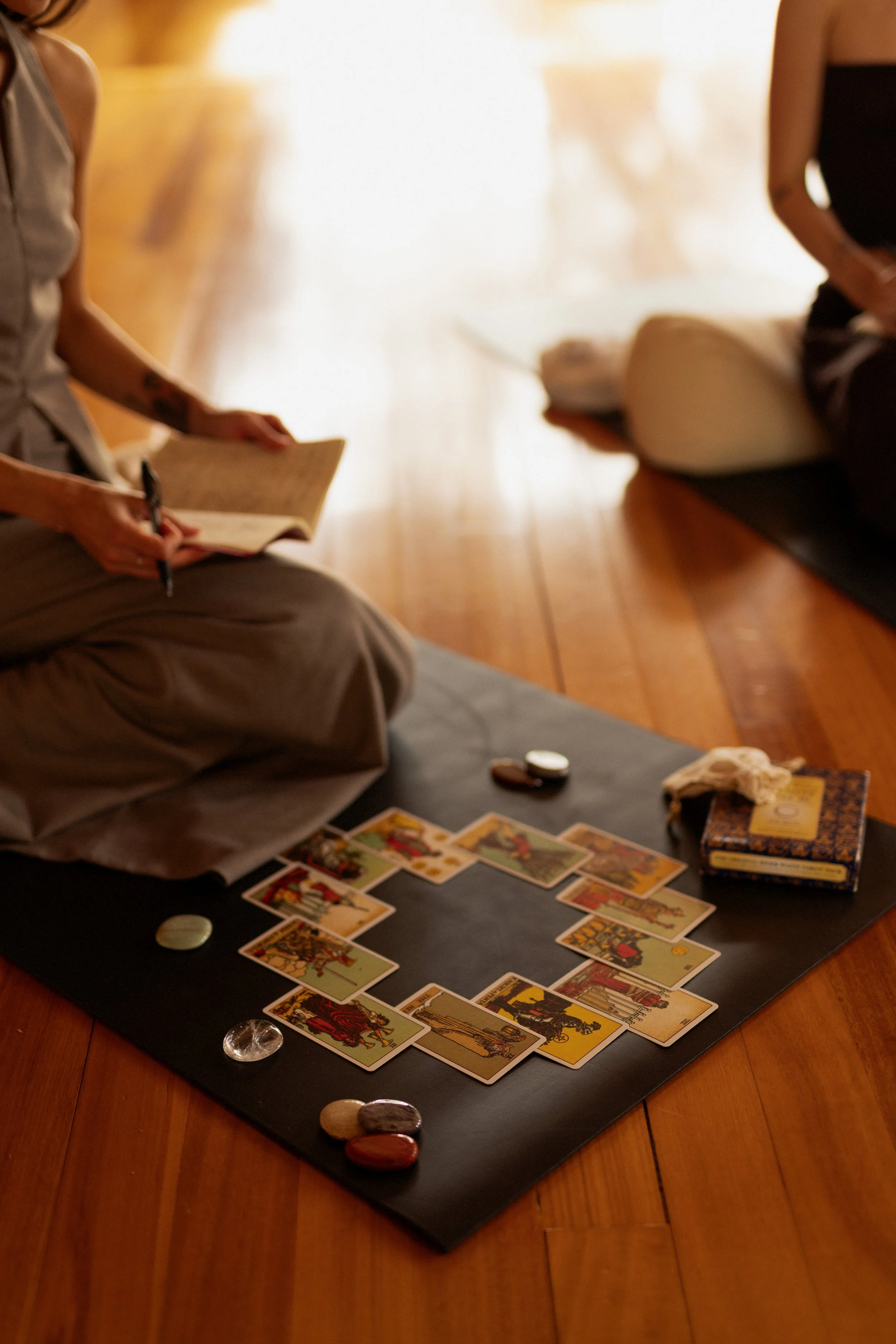 People sitting on cushions, playing a game of tarot cards on a black mat, with tarot decks and stones on the floor, in a warmly lit room.