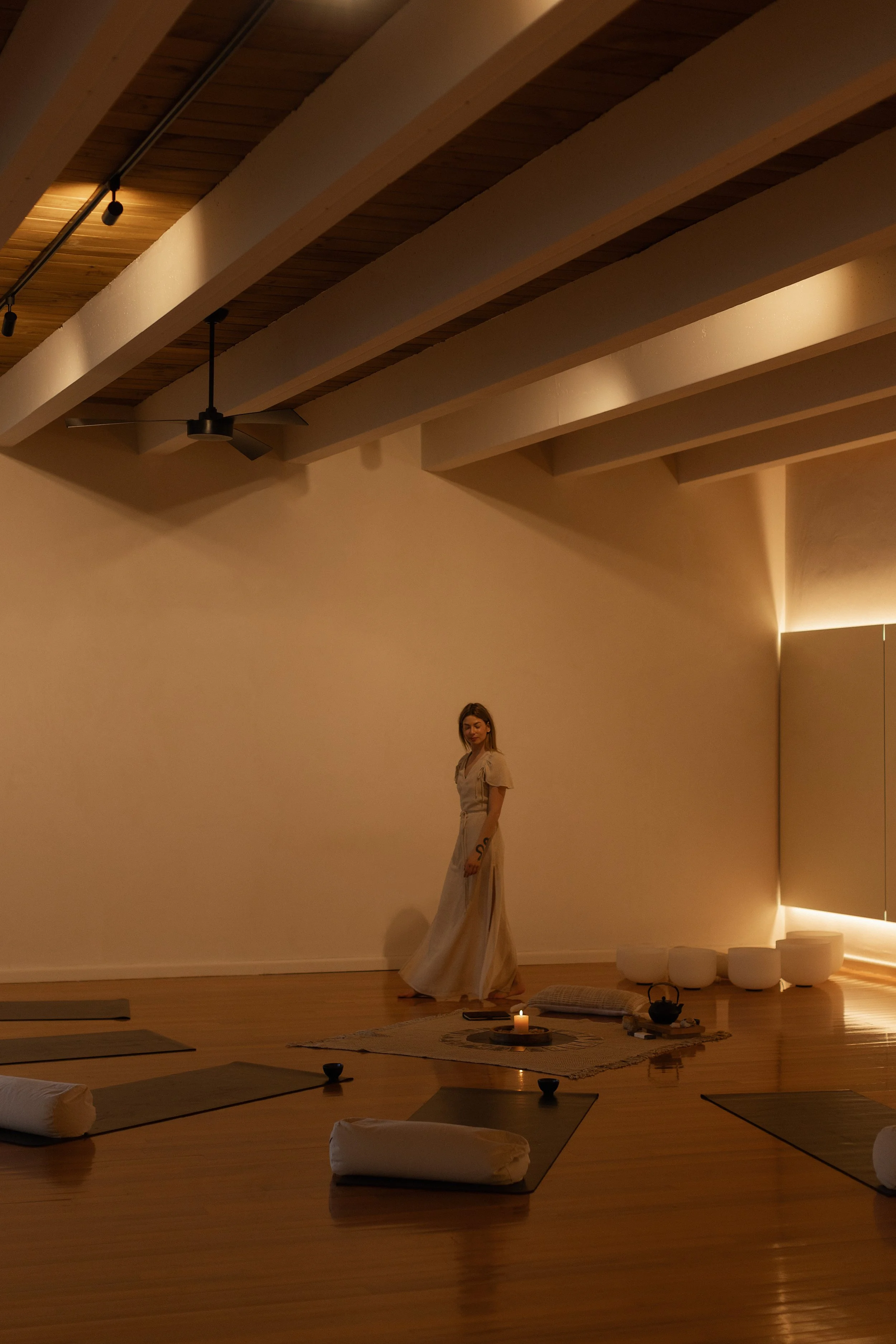 A woman in a long white dress standing in a serene, dimly lit yoga or meditation studio with mats, cushions, a lit candle, and a teapot setup, featuring wooden floors and a ceiling with exposed beams.