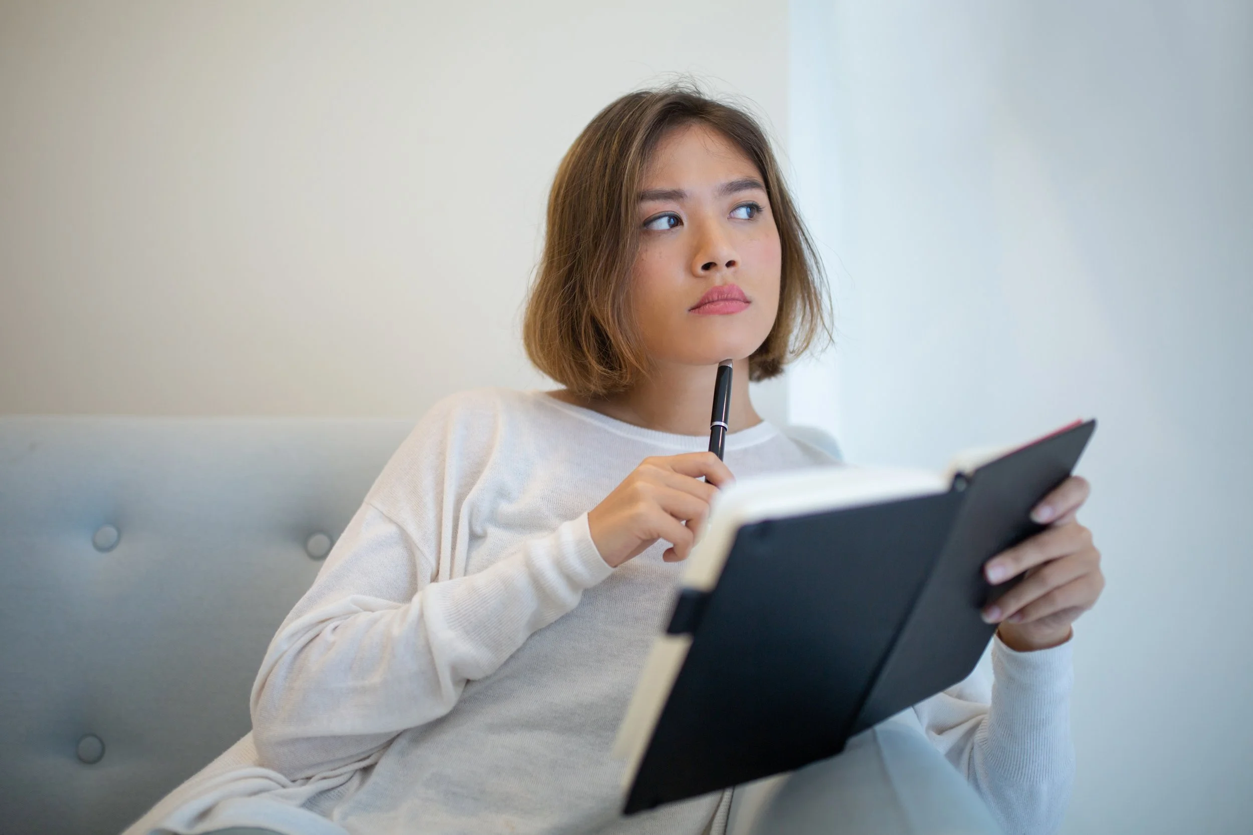 Young woman sitting on a couch with journal and pen, thoughtfully checking in with herself after an online therapy consult session.