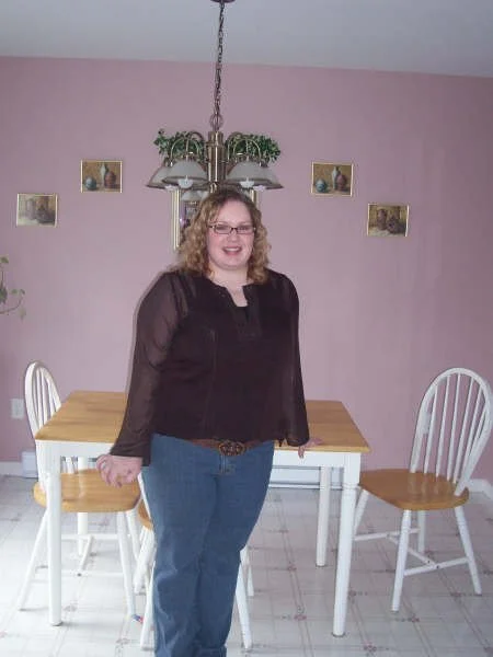 A woman with curly blonde hair and glasses standing in a dining room, smiling. She is wearing a black blouse and blue jeans. Behind her is a wooden dining table with four white chairs and a chandelier hanging above.