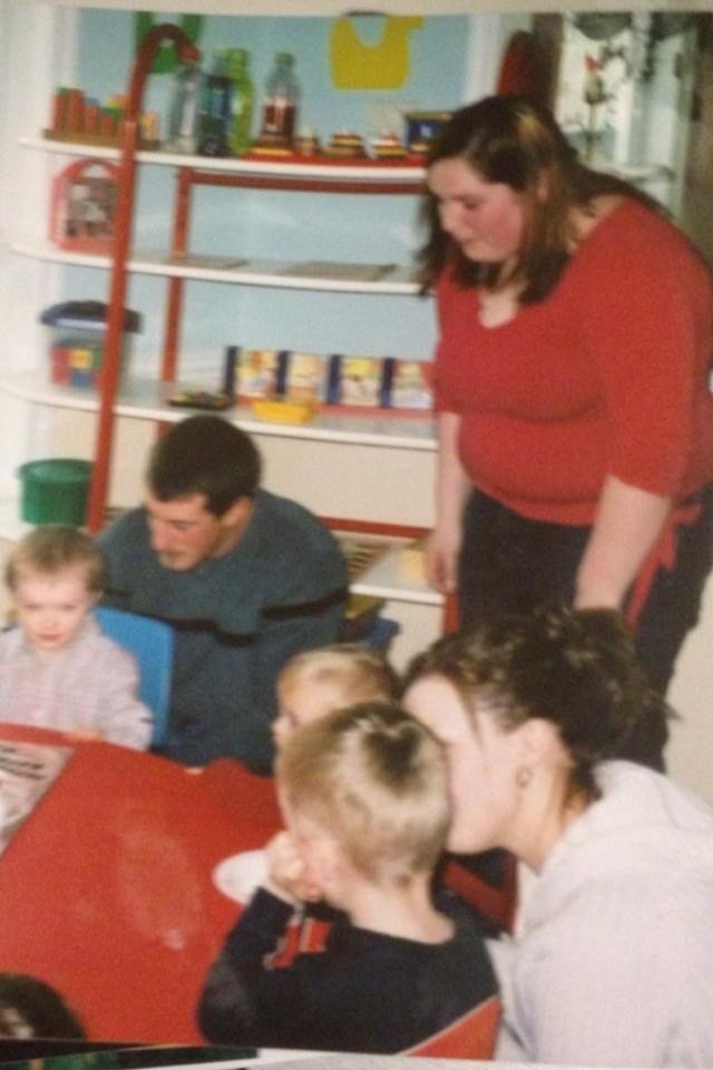 A group of children and adults gathered around a red table, engaging in an activity or conversation in a room with shelves and colorful decorations.