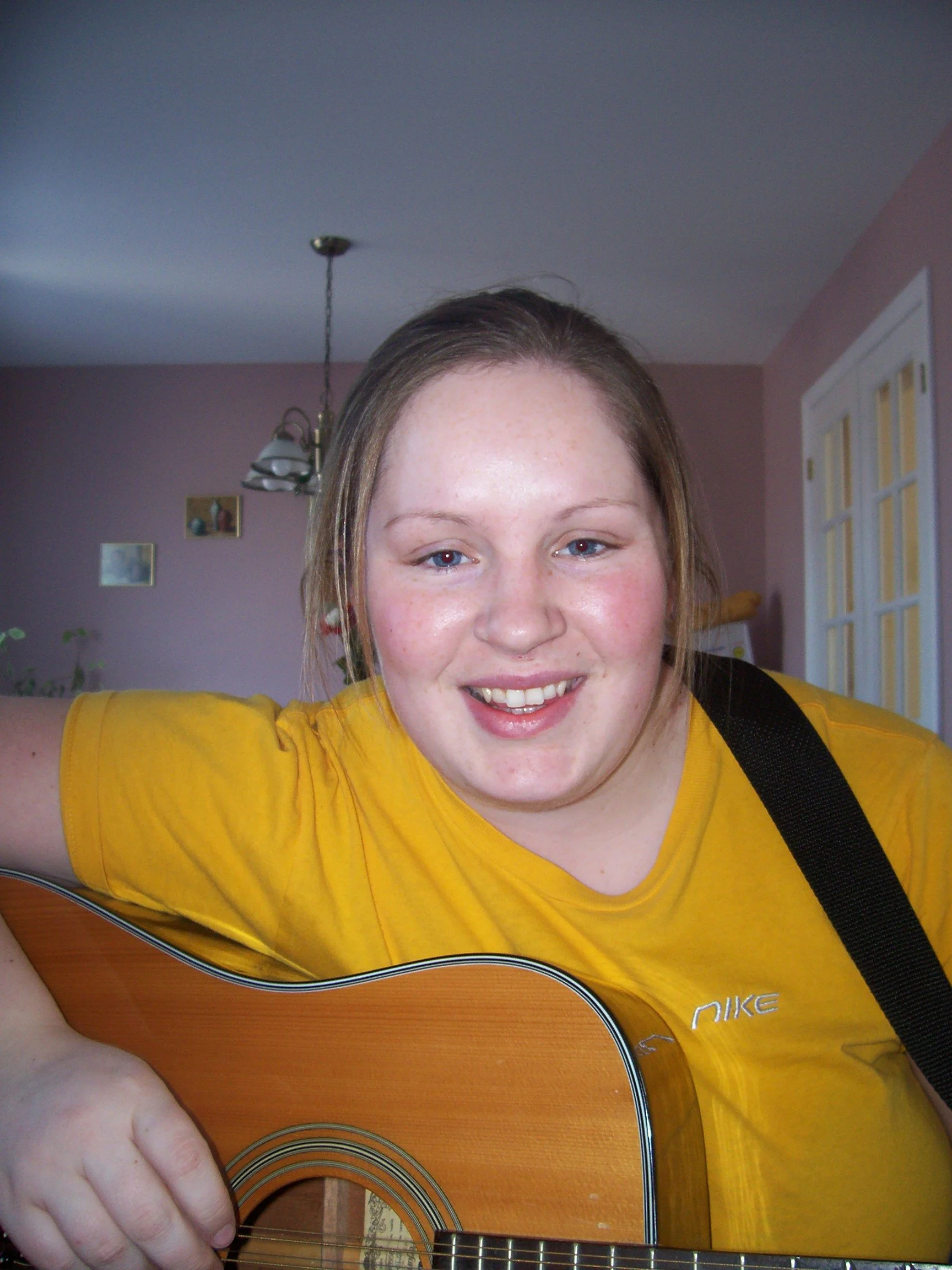 A young woman with fair skin and brown hair playing an acoustic guitar indoors, smiling at the camera, wearing a yellow T-shirt, in a room with pink walls and a white door.