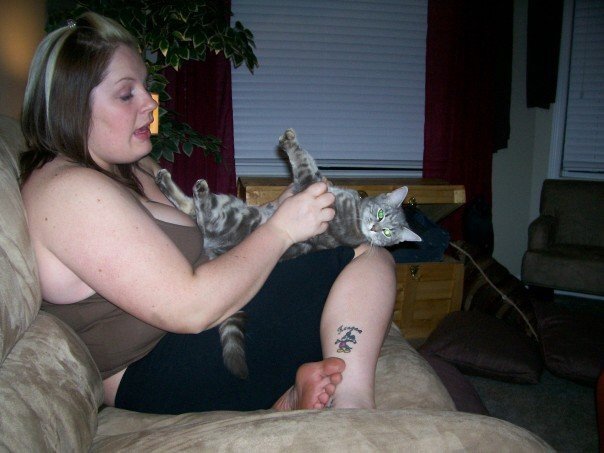 A woman sitting on a beige couch playing with two gray tabby cats indoors at night.