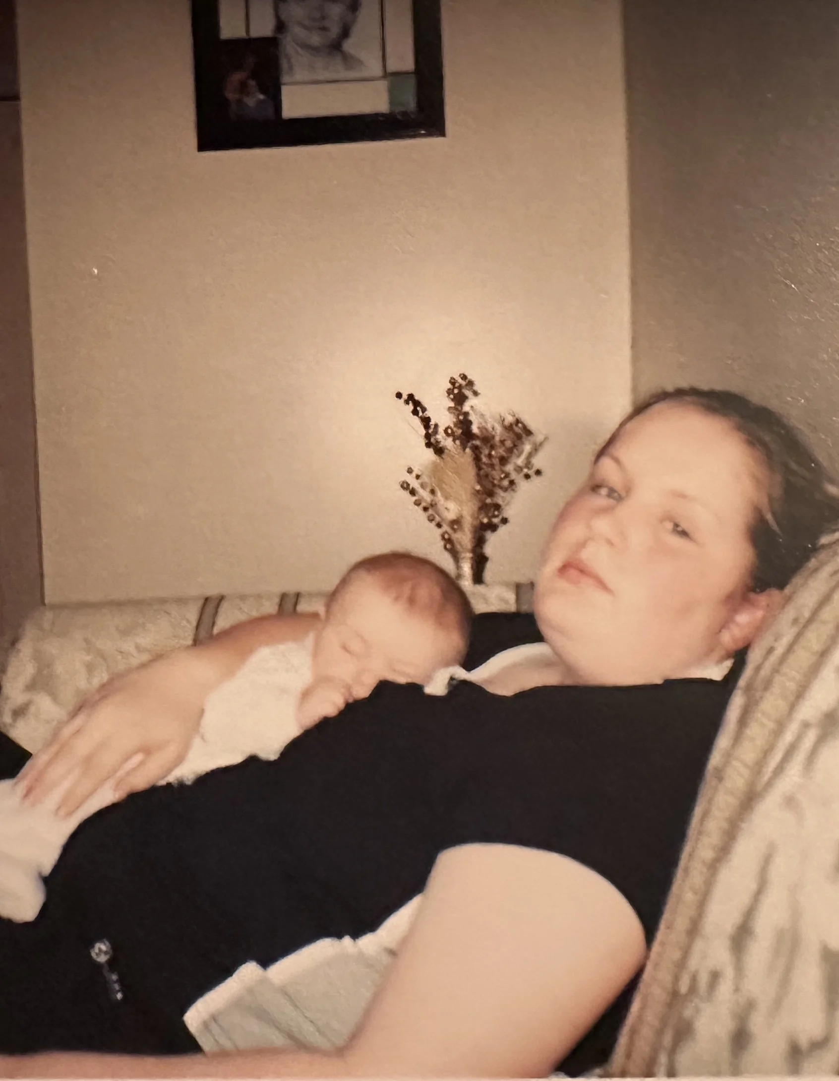 A person with a relaxed expression lying on a couch with a newborn baby resting on their chest, both indoors with a wall and framed photo behind them.