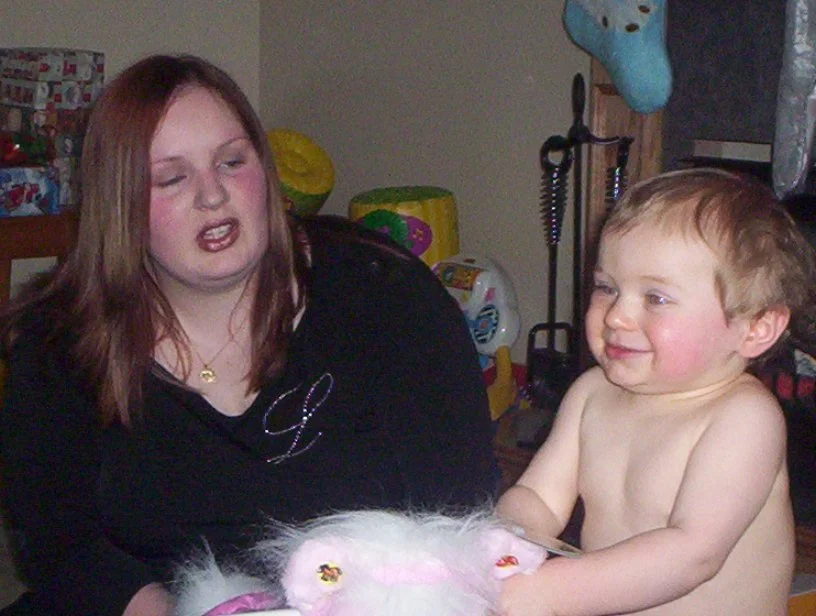 A woman and a shirtless young boy sitting at a table with a white fluffy toy in front of them, in a room with children's toys and decorations in the background.